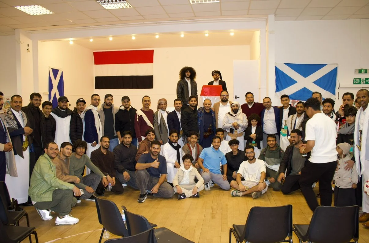 A large group of people, mostly young men, are gathered in a room with flags on the walls, including the flags of Scotland, Egypt, and Scotland. Two men are standing on a table at the back of the room, one speaking into a microphone. The group appears to be celebrating or attending a cultural event.