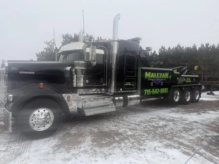 A black semi-truck with green and yellow graphics and text, parked on a snowy ground with a forest in the background.