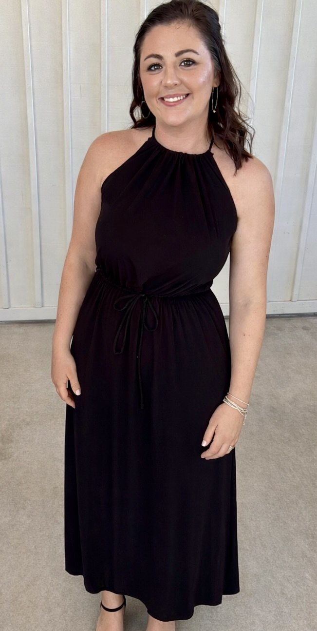 A woman with dark brown hair, wearing a black sleeveless dress with a high neckline, standing in front of a white wooden wall. She is smiling, wearing hoop earrings and bracelets, and black ankle-strap shoes.