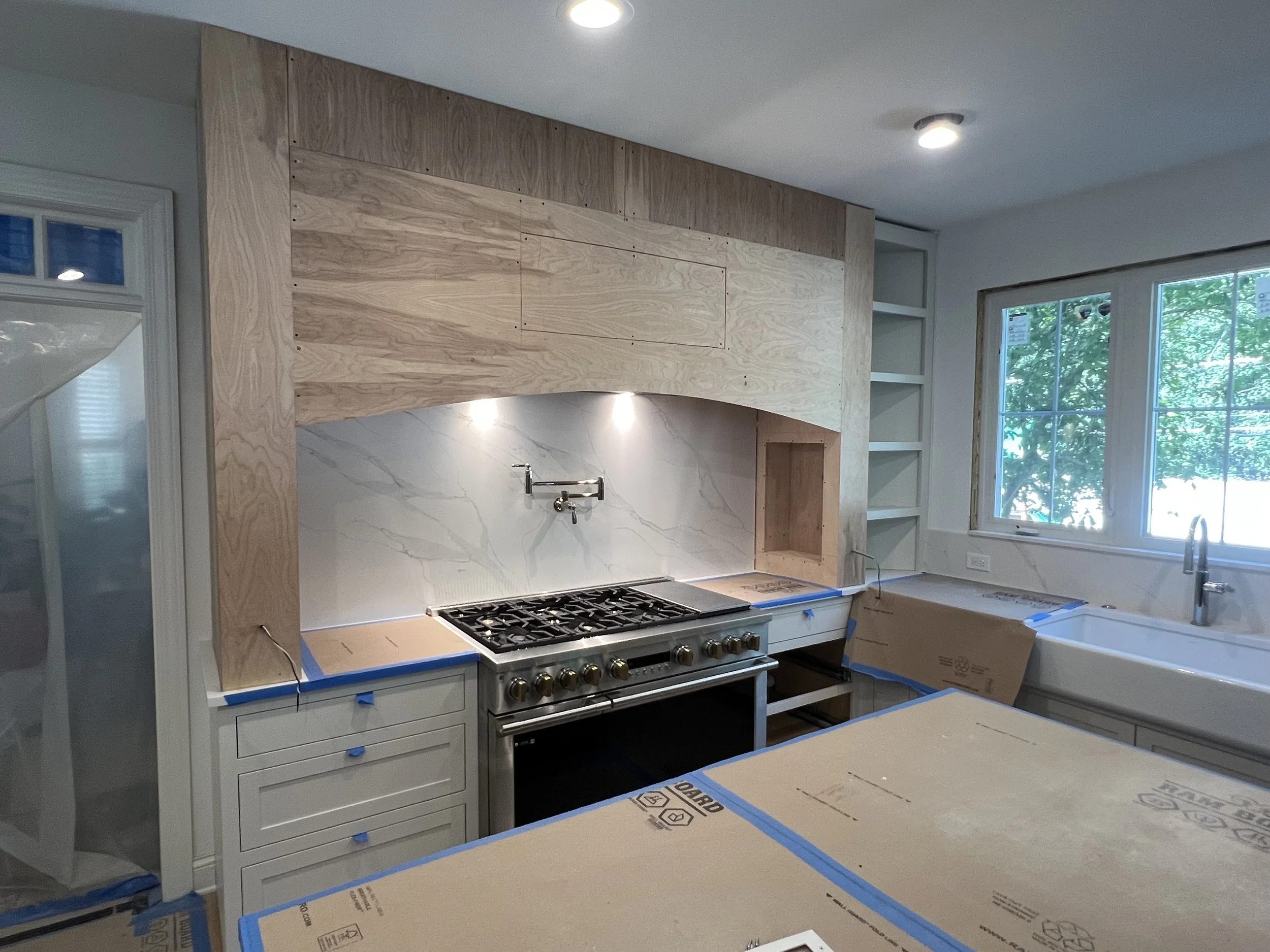 Kitchen under construction with uninstalled cabinets, a stove, and a large window showing trees outside.