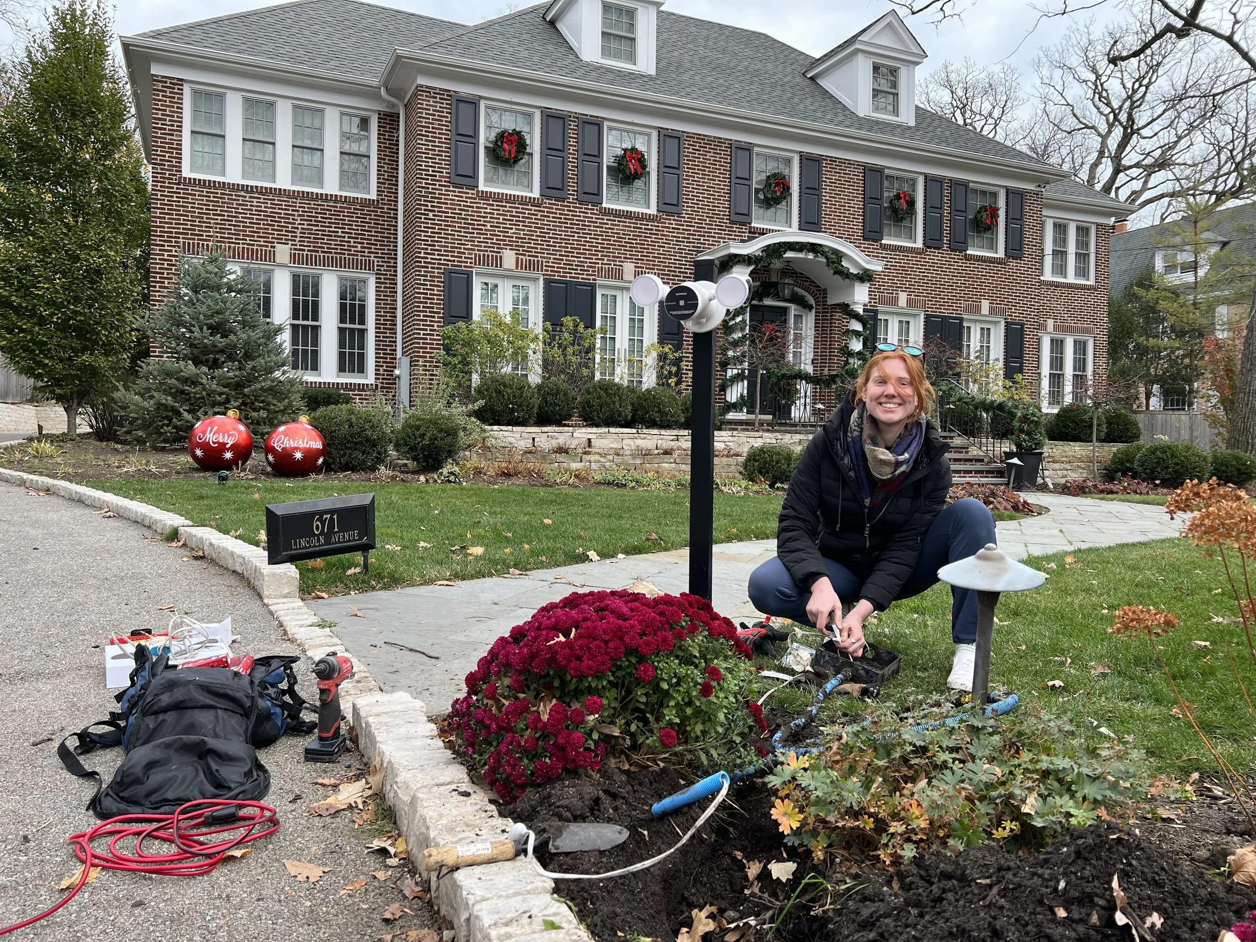 A woman crouching in a front yard garden of a large brick house with Christmas decorations, installing outdoor lighting. The garden has flowers, a small lamp, and Christmas ornaments. There are tools and a backpack on the ground nearby.