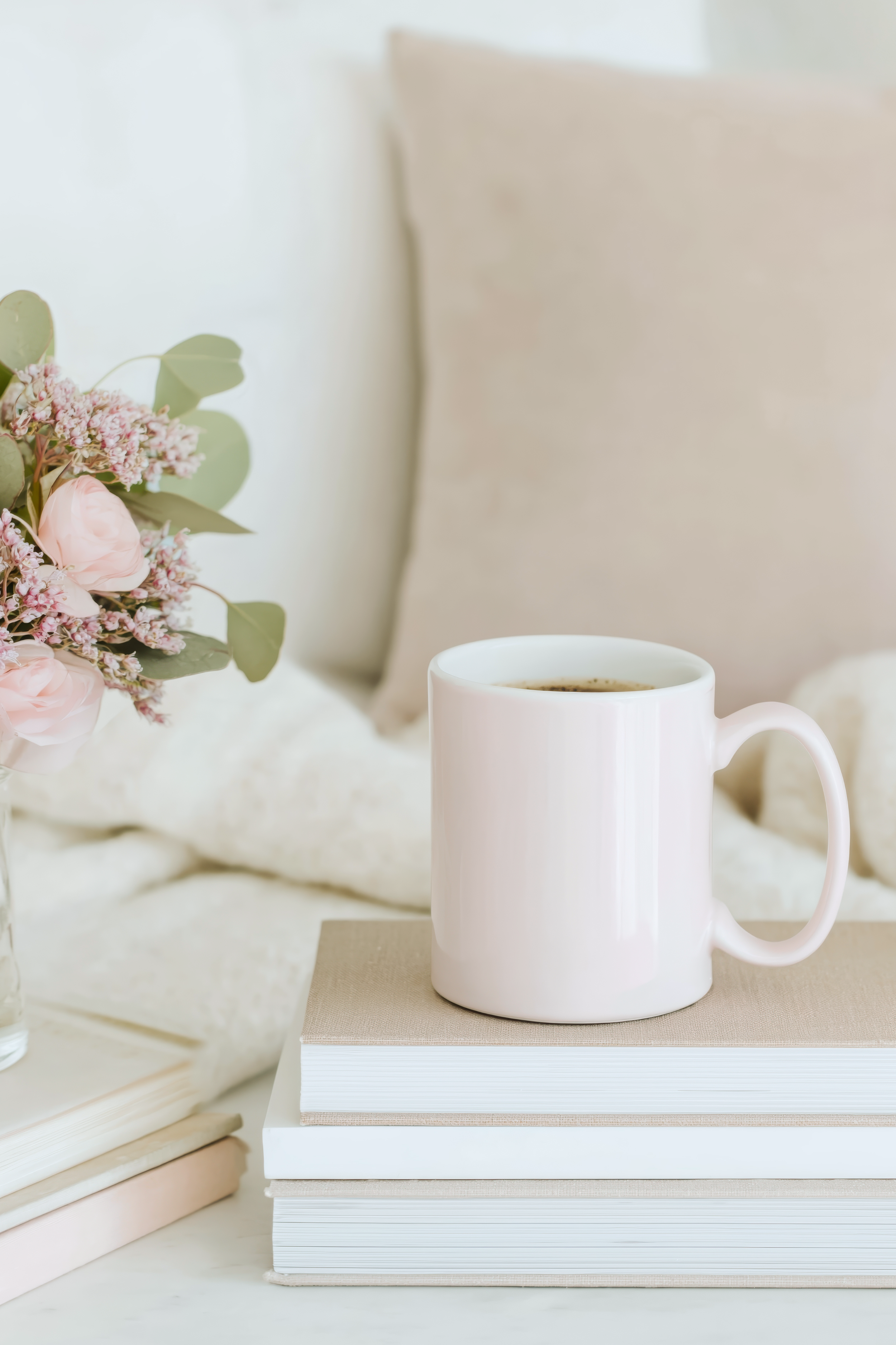 A white coffee mug filled with coffee placed on top of three stacked books on a light surface, with a pink flower bouquet and a beige pillow in the background.