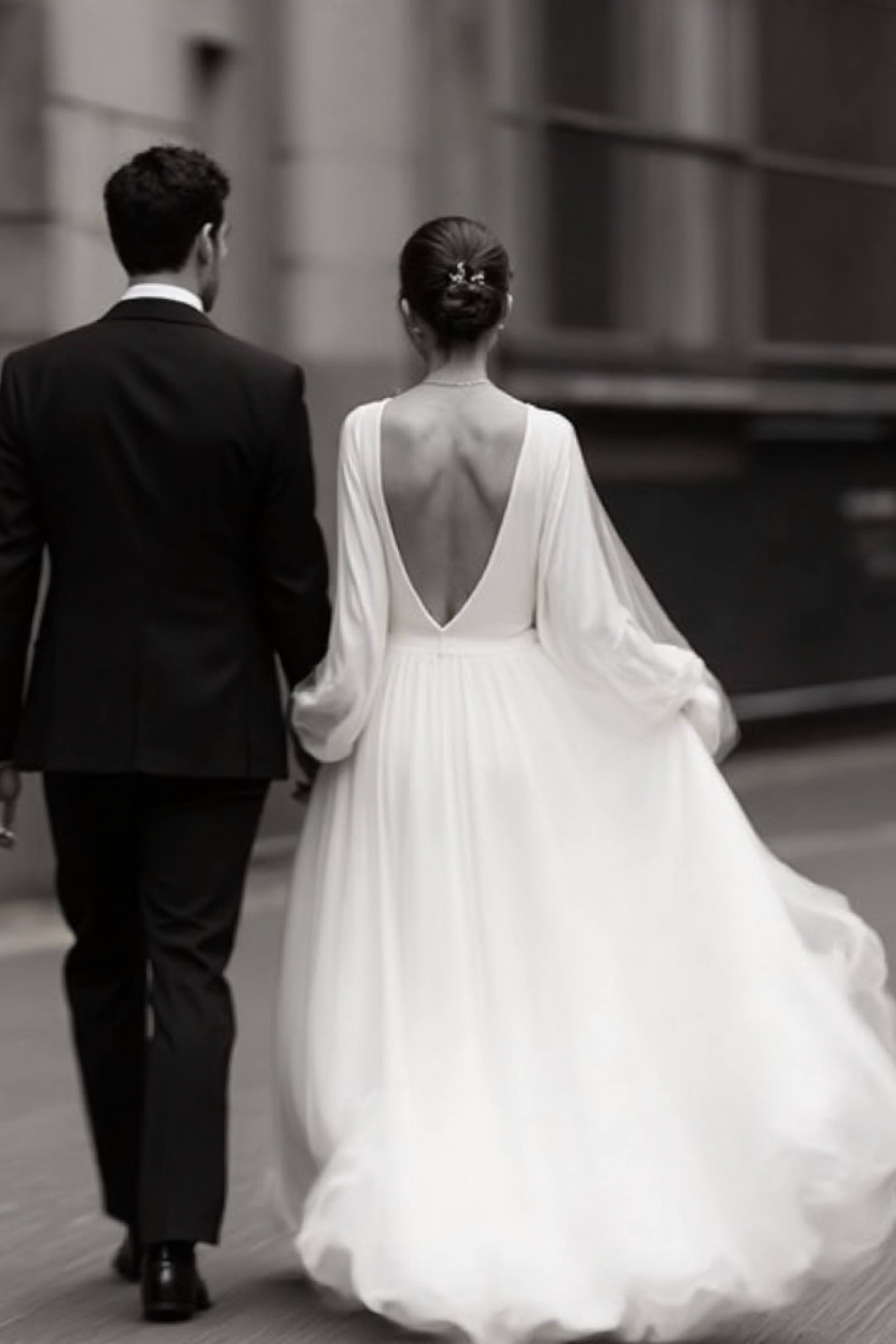 A black-and-white photo of a couple walking away, the man wearing a tuxedo and the woman in a long, elegant wedding dress with an open back.