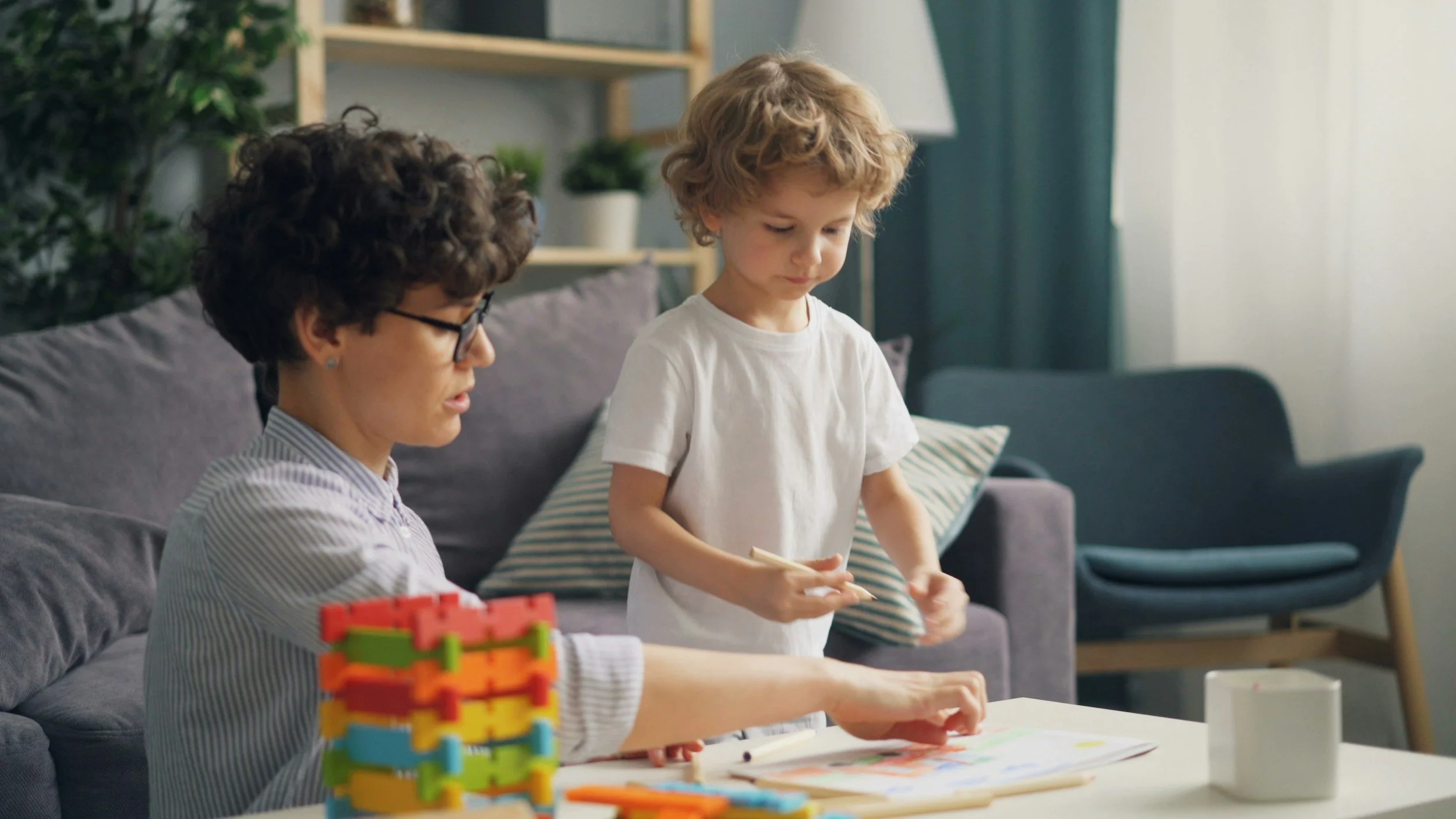 A woman and a young boy sitting at a table engaging in an activity, with toys and craft supplies on the table. The woman has short curly hair and glasses, and the boy has blond curly hair, wearing a white t-shirt. The background shows a couch, pillow, and a bookshelf with plants.