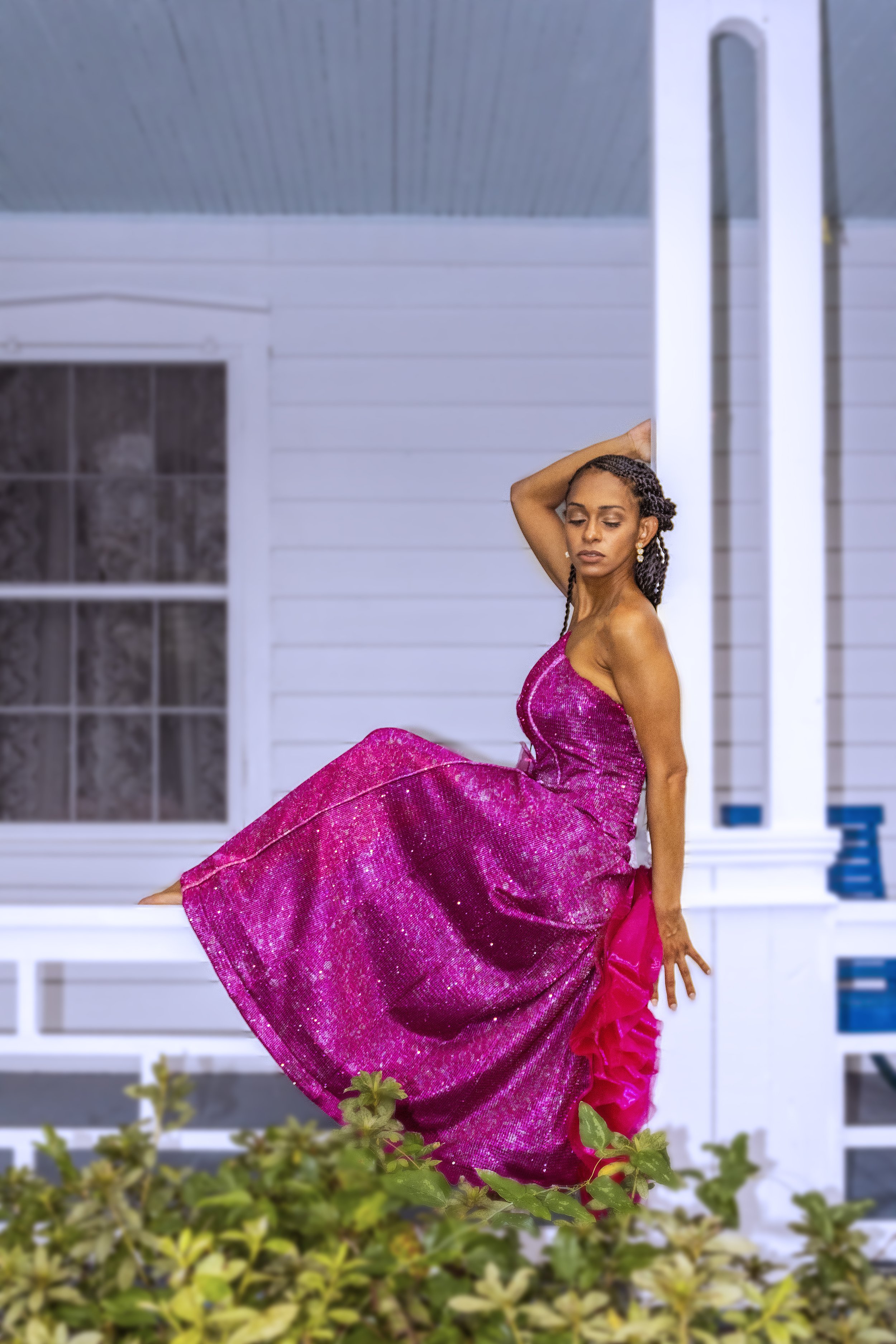 A woman in a shiny pink dress sits gracefully on the edge of a white porch or deck, with her eyes closed and one arm resting on her head, surrounded by greenery with a white house in the background.