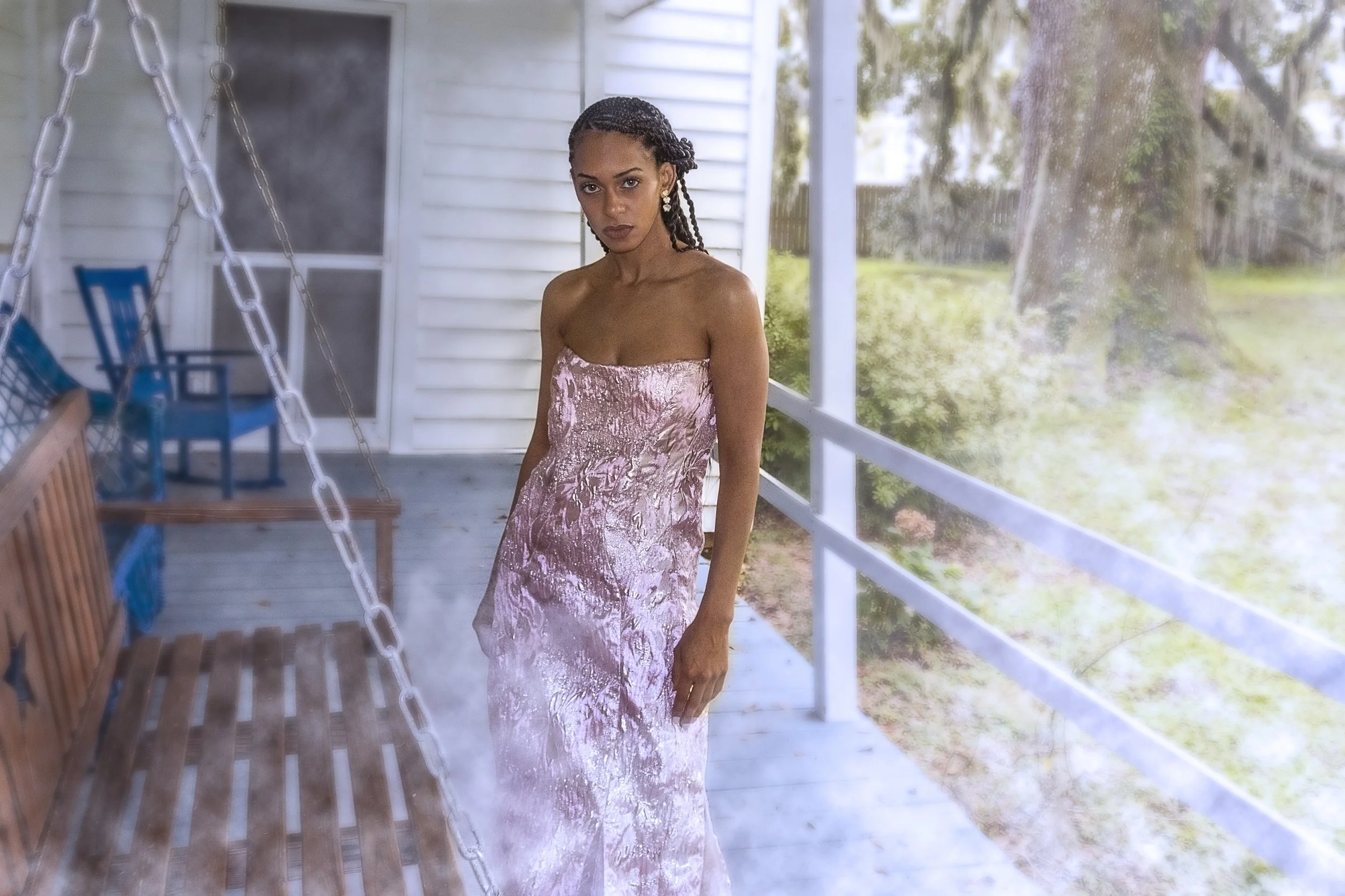 A woman in a pink velvet strapless dress standing on a porch next to a glass screen door, with trees and greenery visible outside. Fashion Photography.