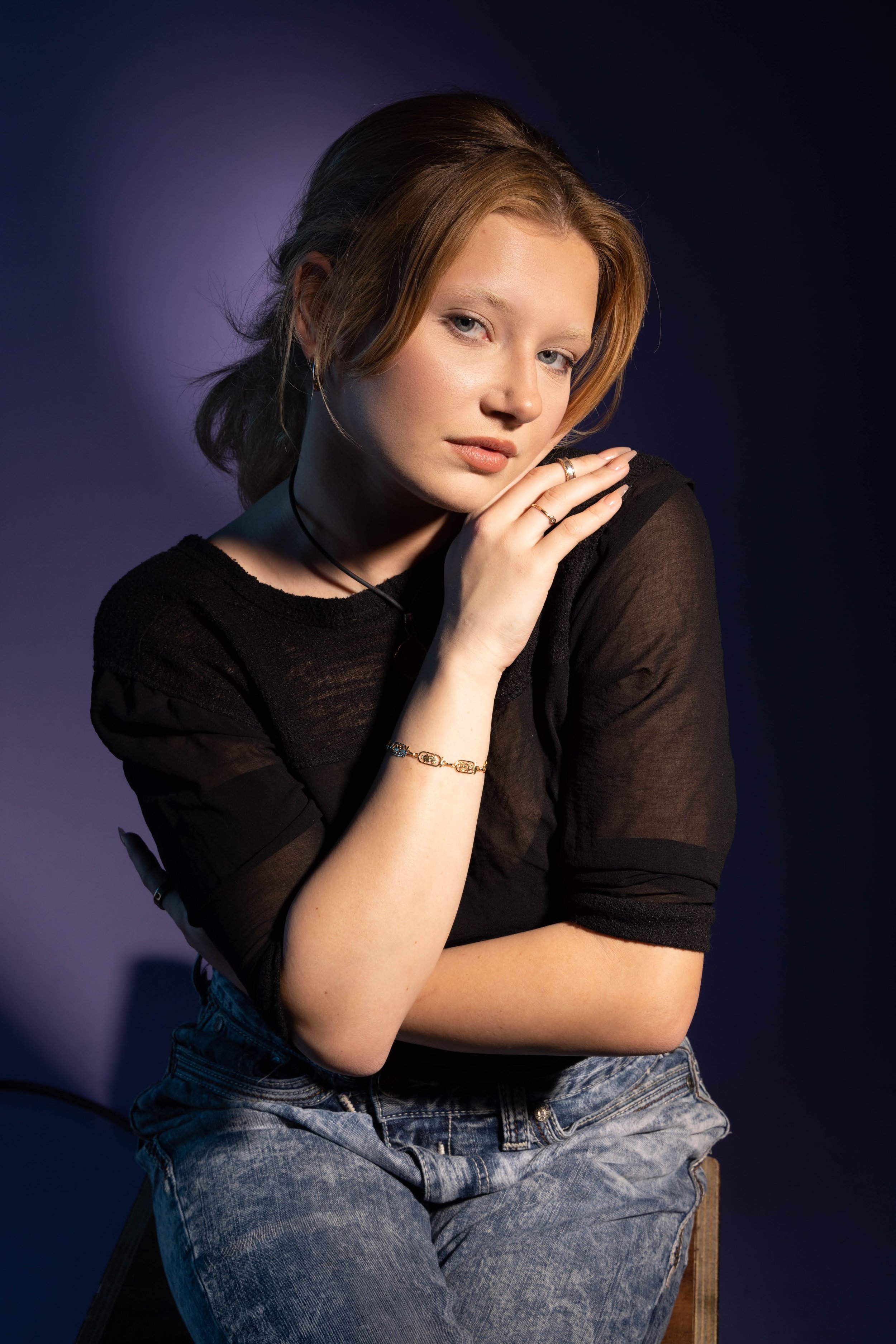 A young woman with shoulder-length red hair, wearing a black sheer top and blue jeans, posing with her left hand gently touching her chin against a dark background with purple lighting.