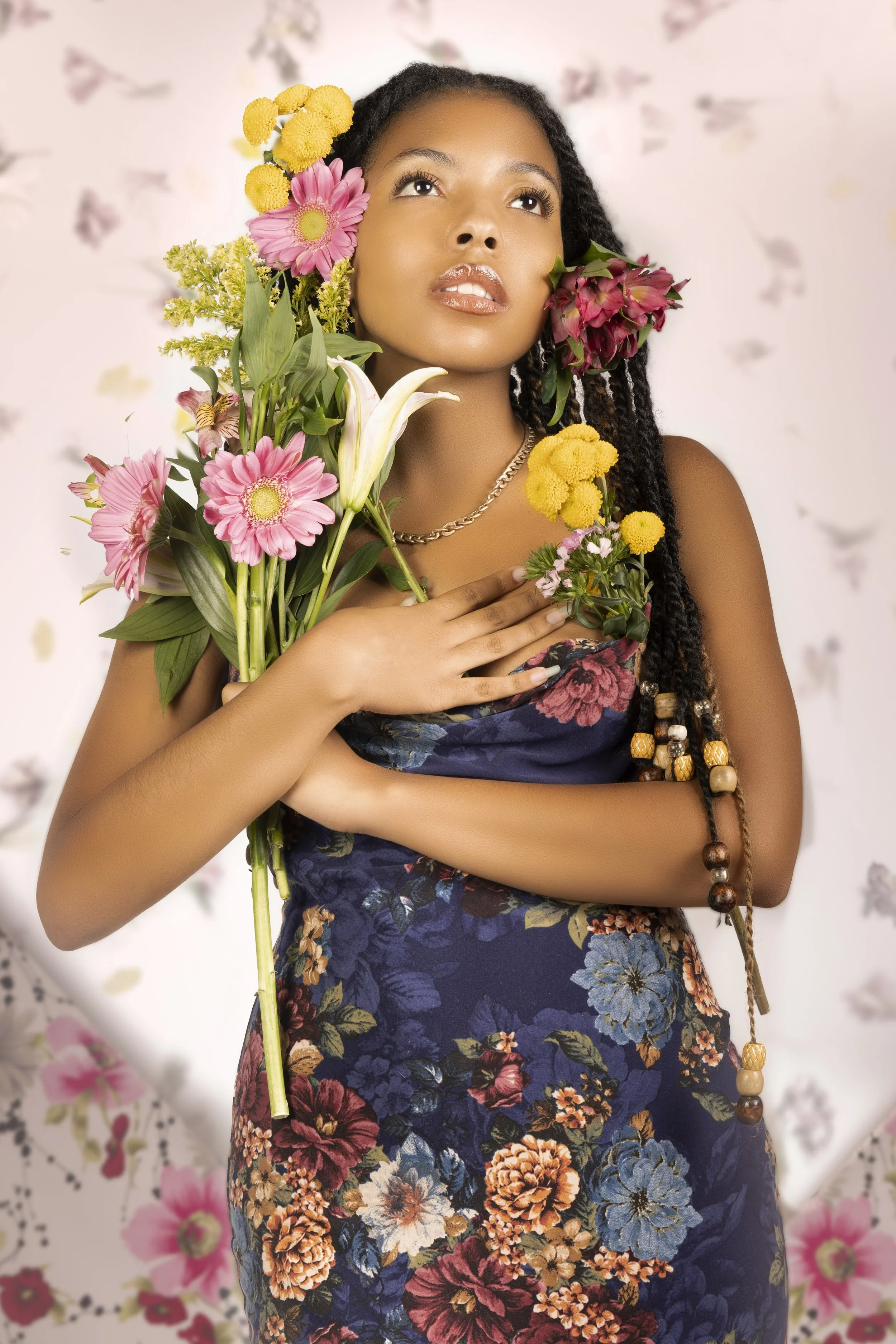 A woman with long, braided hair holding a bouquet of colorful flowers near her chest, looking upward. She is dressed in a floral dress and has flowers in her hair, with a softly blurred background featuring pastel flowers.
