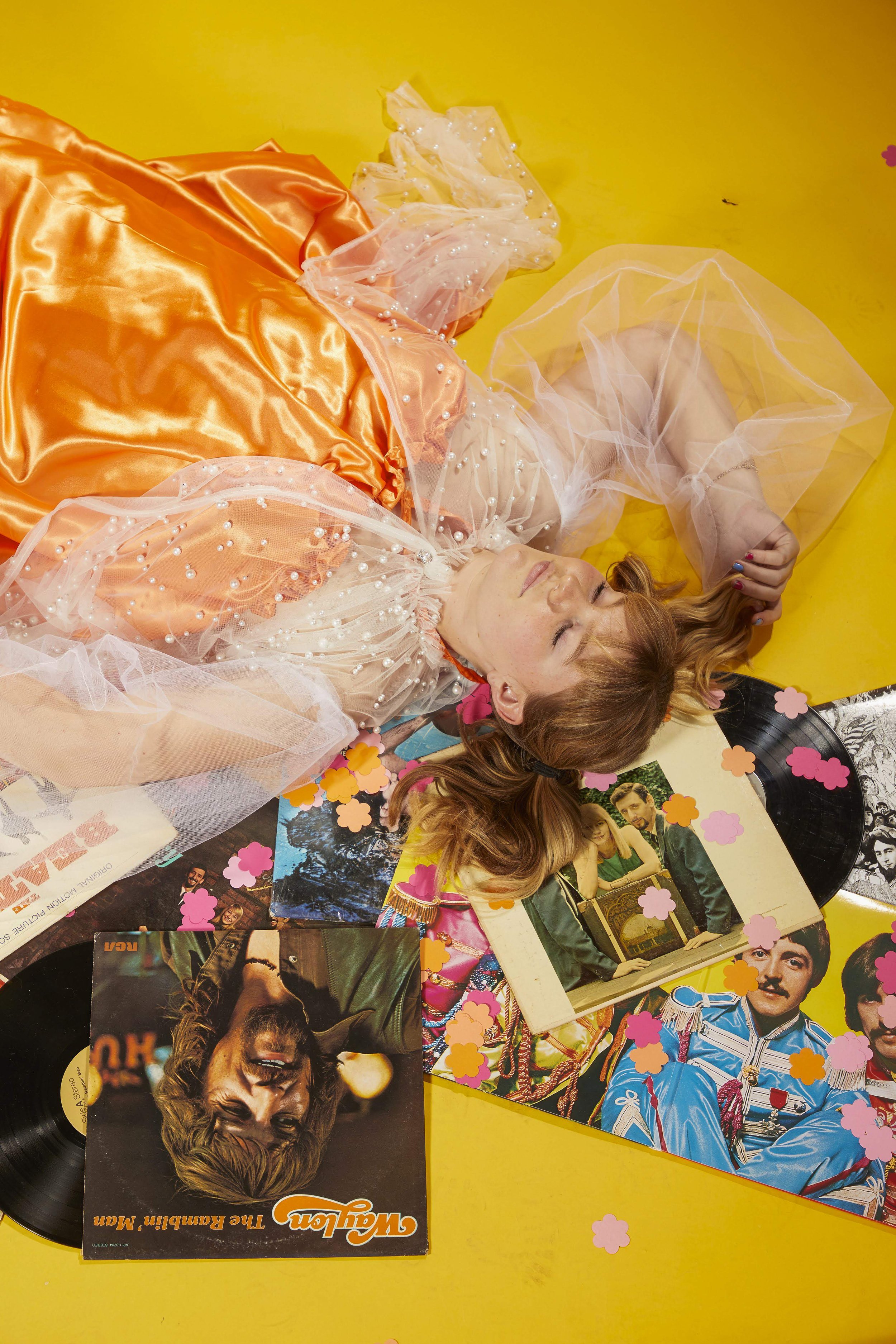 A woman with red hair and closed eyes lying on a yellow floor, surrounded by vintage vinyl records, colorful photos, and pink and orange flower confetti.
