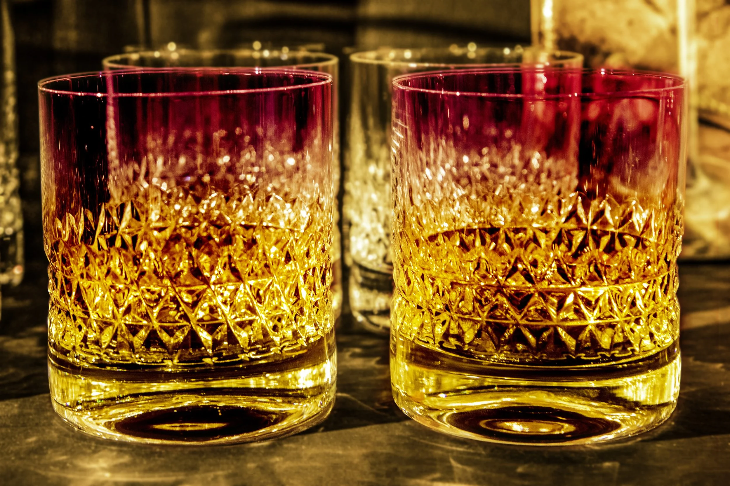 Two glasses of whiskey with intricate diamond cut patterns, illuminated with warm orange and purple light, placed on a dark wooden surface.