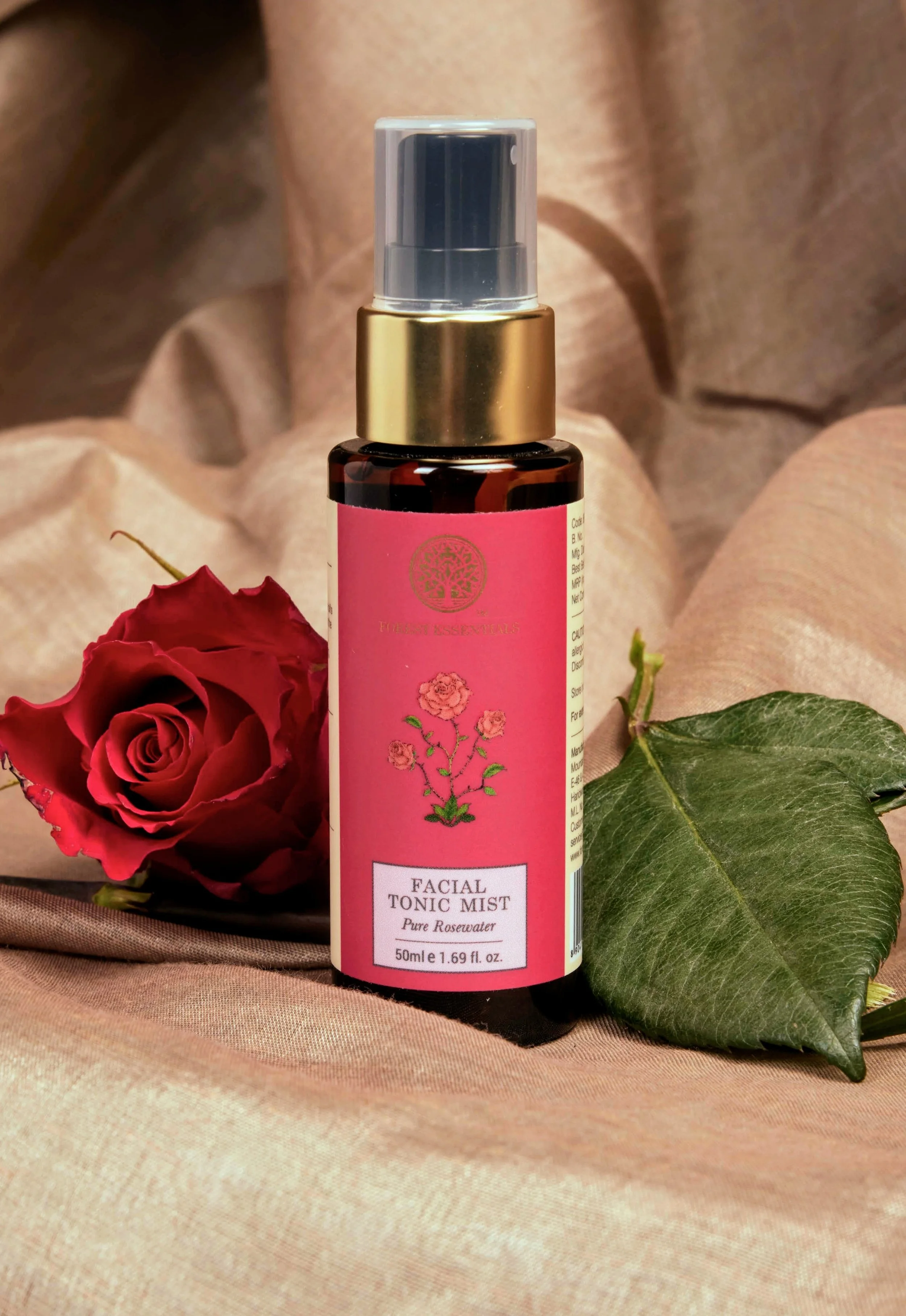 A pink bottle of facial tonic mist with a floral illustration and gold cap, placed beside a red rose and green leaf on a soft fabric background.