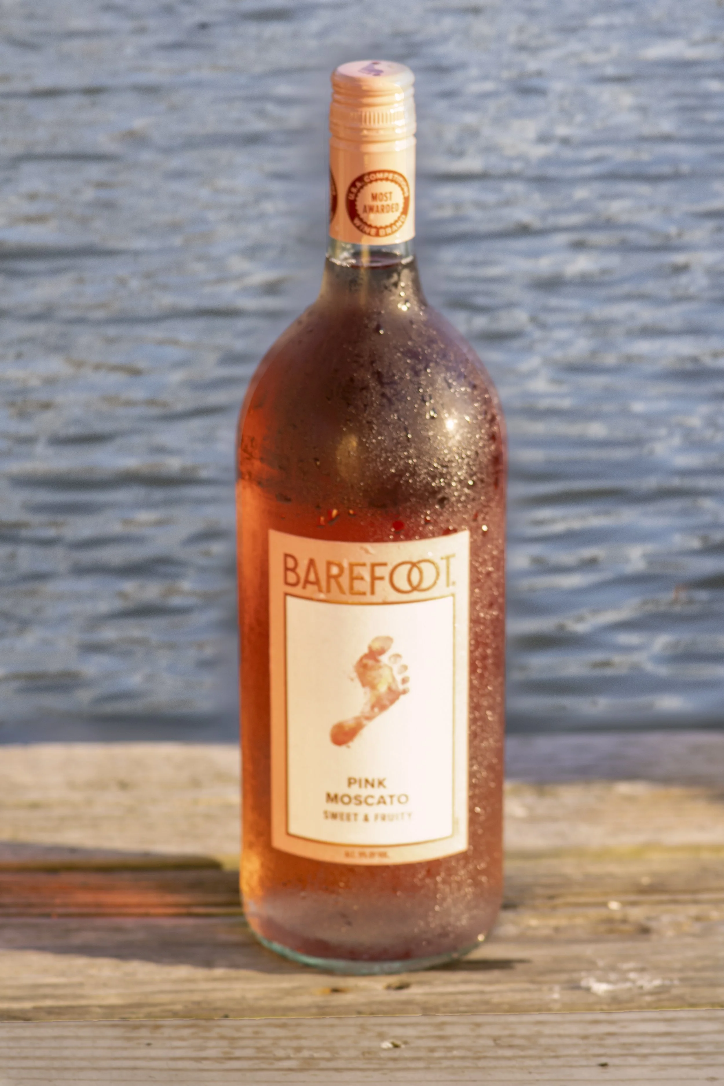 A bottle of Barefoot Pink Moscato wine with condensation on it, placed on a wooden surface by a body of water.