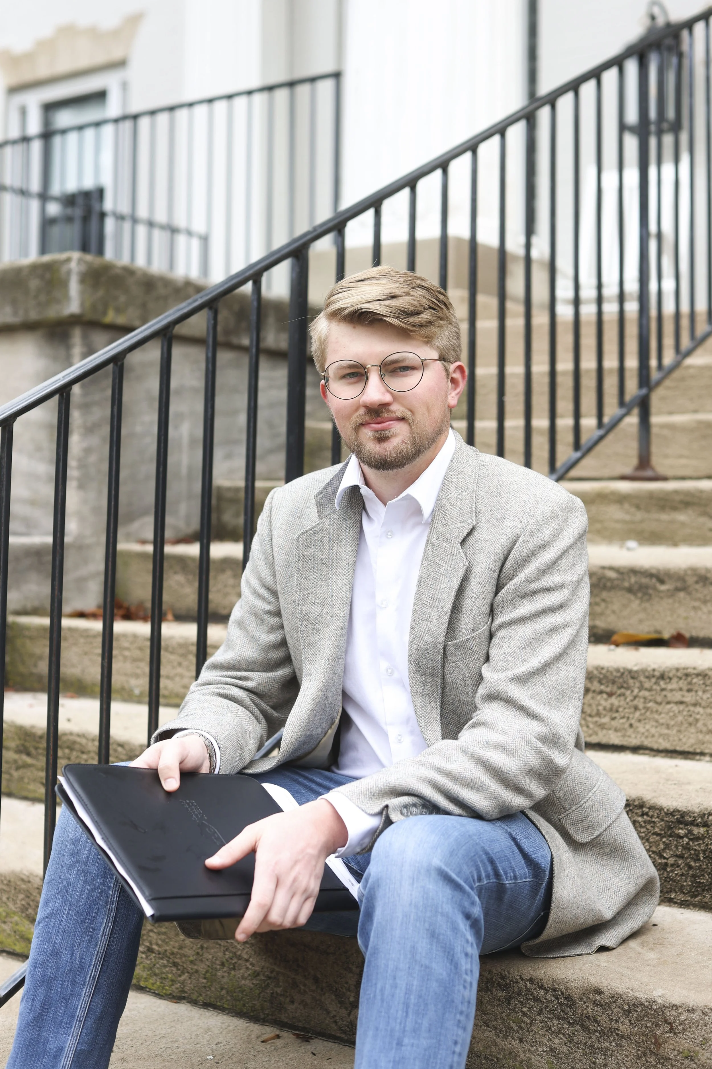 Tate Ohmer sitting on outdoor stairs, holding a black folder, with a building and railing in the background.