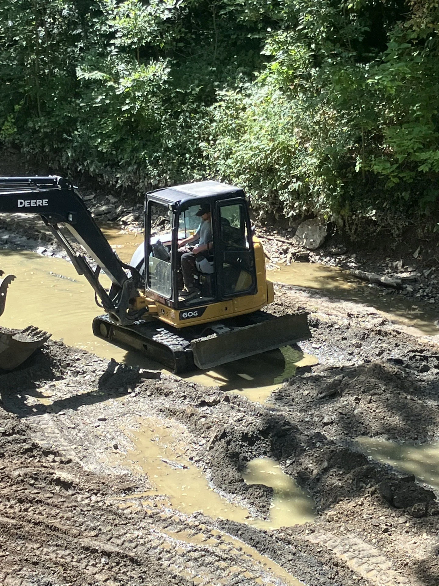 A small yellow and black excavator working in a muddy construction site surrounded by dirt, rocks, and green foliage.