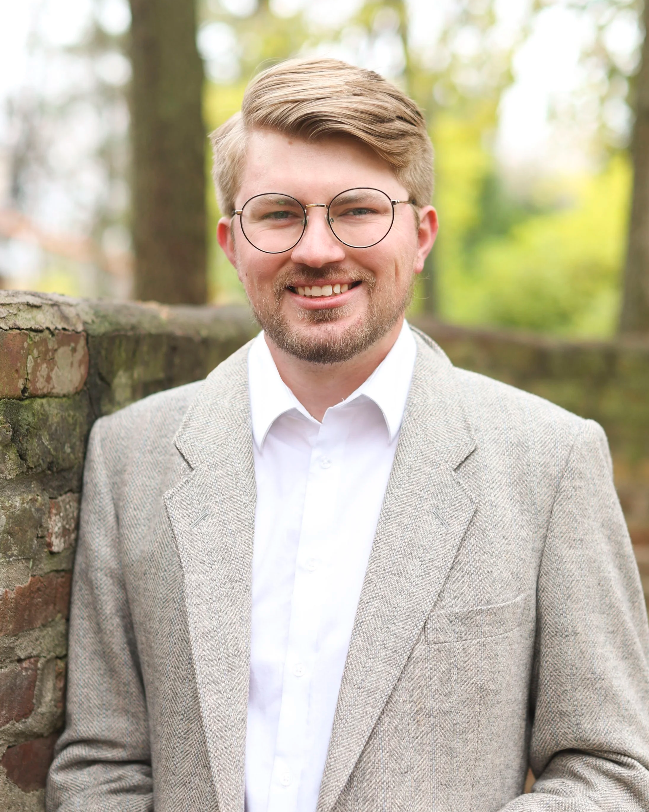 A young man with blonde hair, glasses, and a beard, smiling, wearing a light gray blazer and white shirt, leaning against a brick wall outdoors with trees in the background.