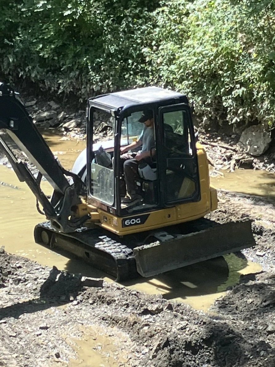 Tate Ohmer, dredging a creek, operating a small yellow and black excavator on a muddy construction site surrounded by green trees.