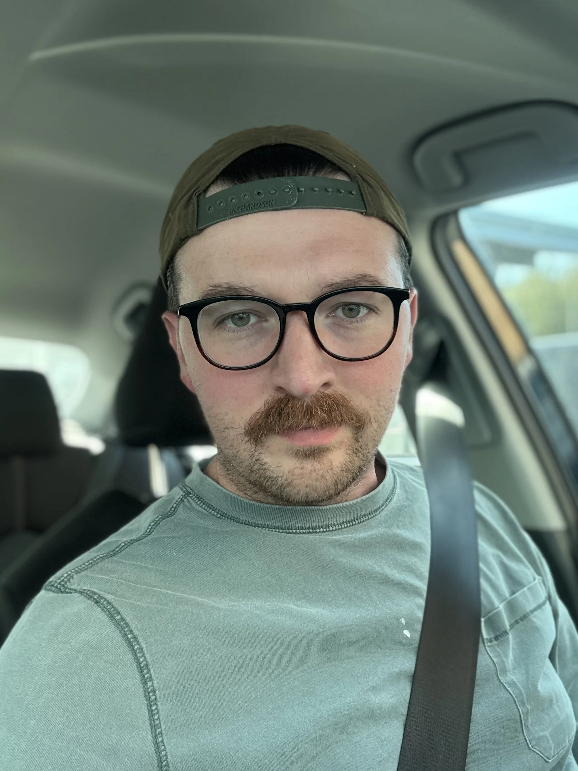 A man with glasses, a mustache, and a beard sitting in a car, wearing a green cap backward and a light green shirt.
