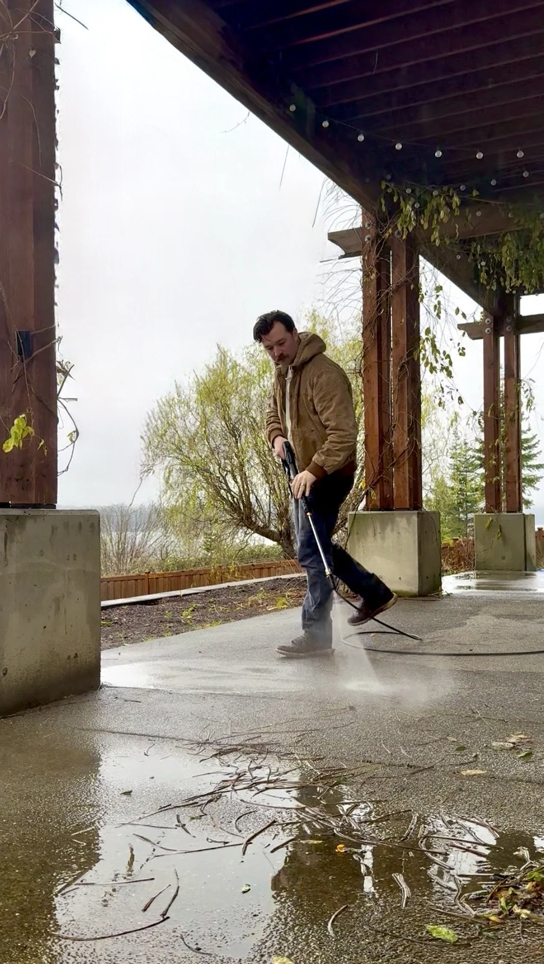 A man wearing a brown jacket and black pants pressure washing a concrete patio under a wooden pergola on a cloudy day.