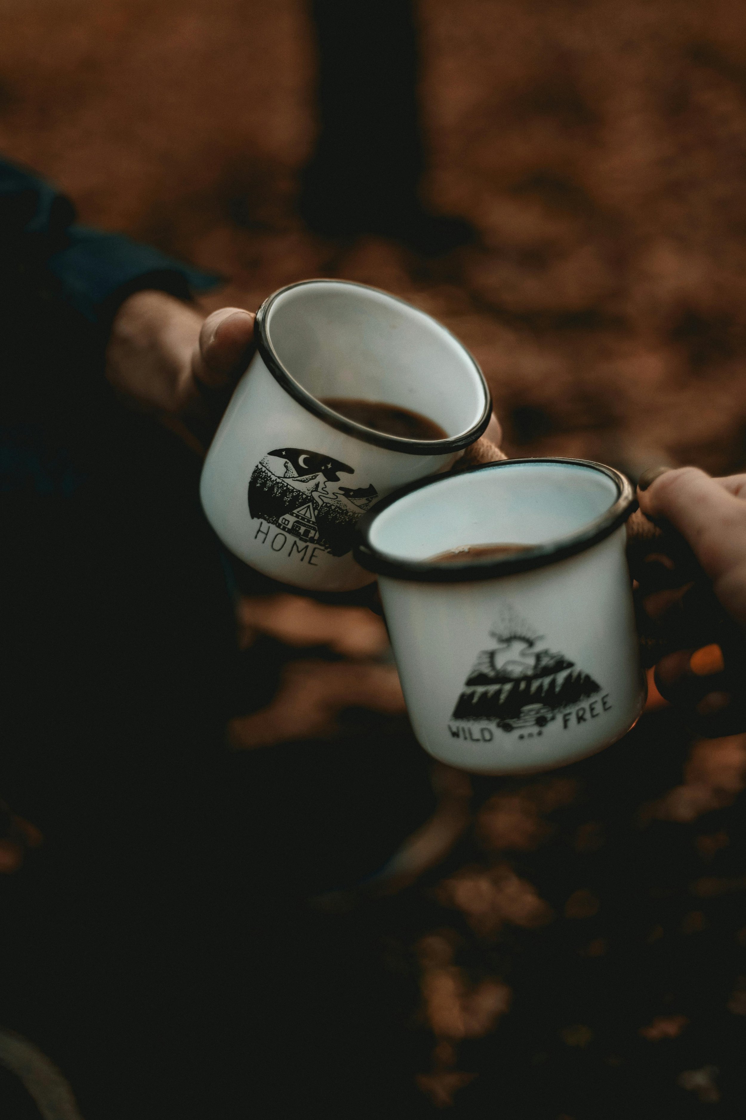 Two hands holding white enamel mugs with black rims, one with a 'Home' design and the other with a 'Wild and Free' design, filled with coffee, against a blurred autumn leaves background.