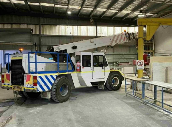 An indoor industrial facility with a large white and blue mobile crane parked inside. The crane has an extendable arm and is marked with yellow and black caution tags, including an oversize warning sign.