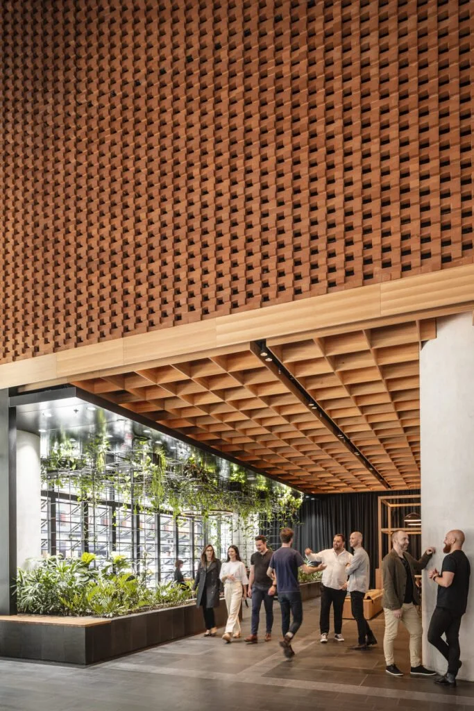 Group of people socializing and walking inside a modern, stylish indoor space with large windows, green plants, and a decorative wooden ceiling.
