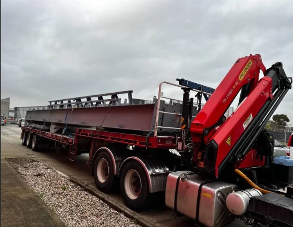 A flatbed truck with a mounted crane parked on a gravel and pavement surface, overcast sky in the background.