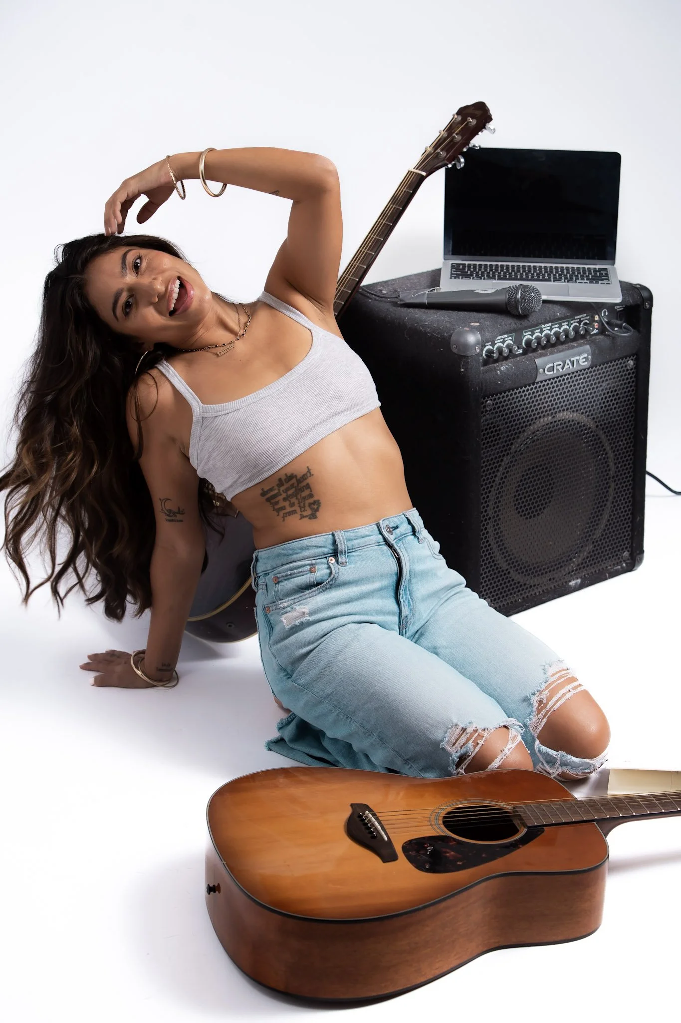 A young woman with long hair, tattoos, and casual clothing, smiling and posing on the floor with musical instruments and equipment around her, including an acoustic guitar, microphone, amplifier, and a laptop.