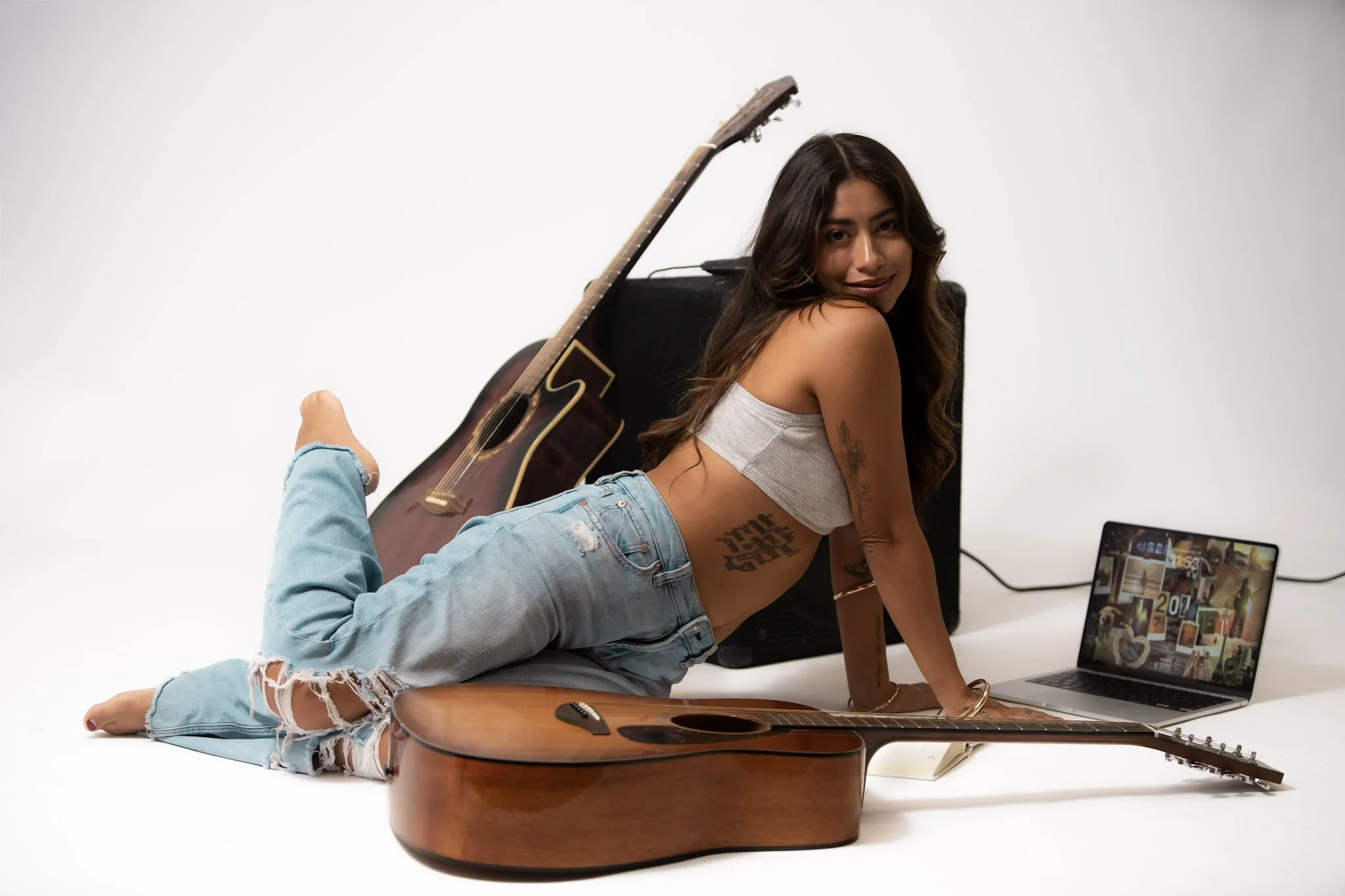 Young woman lying on the floor with a guitar, laptop, and speaker, smiling at the camera.
