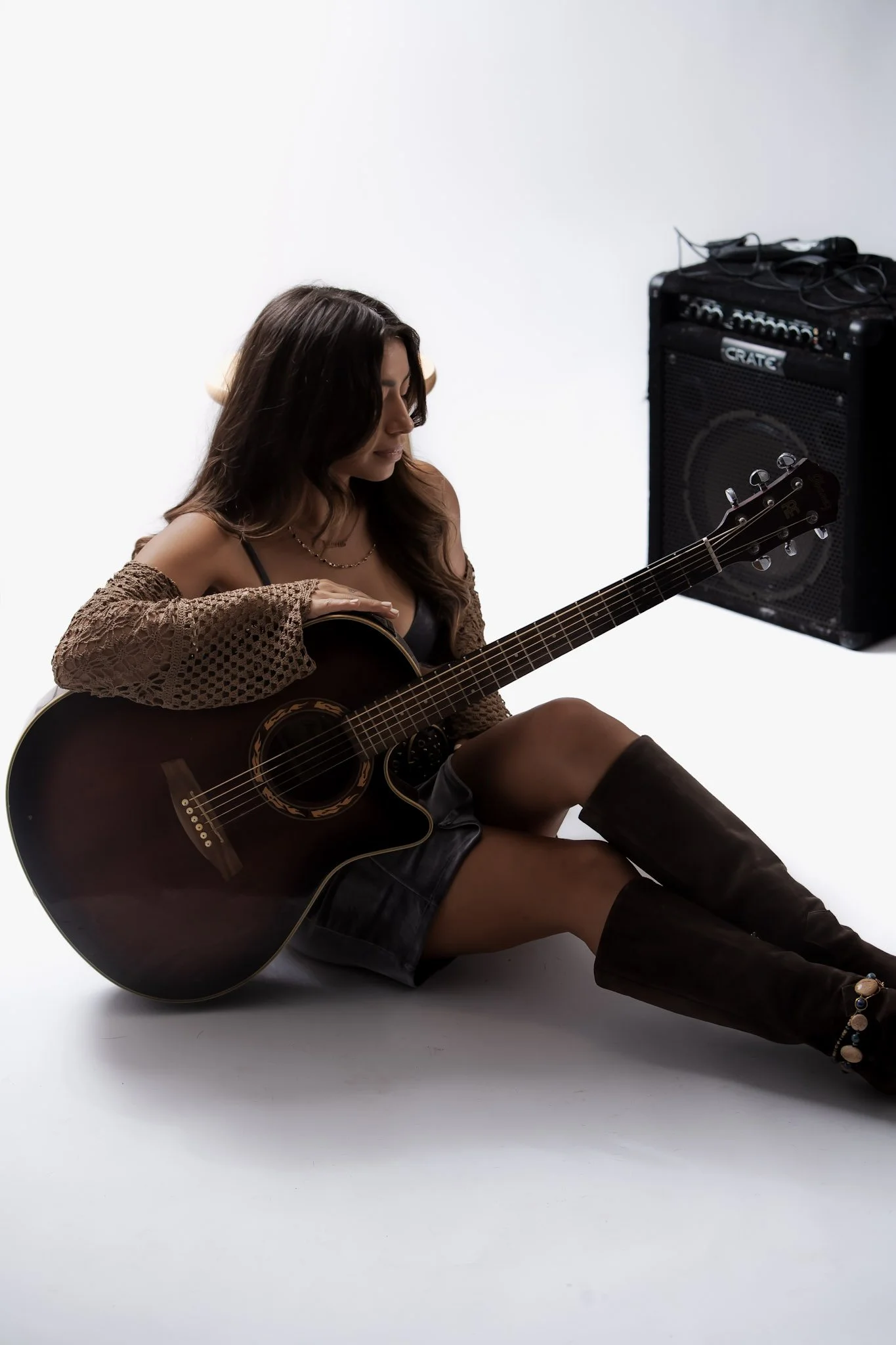 A woman sitting on the floor playing an acoustic guitar with an amplifier behind her against a plain background. Songwriters of Oahu. Country Music on Oahu. Blues music on Oahu