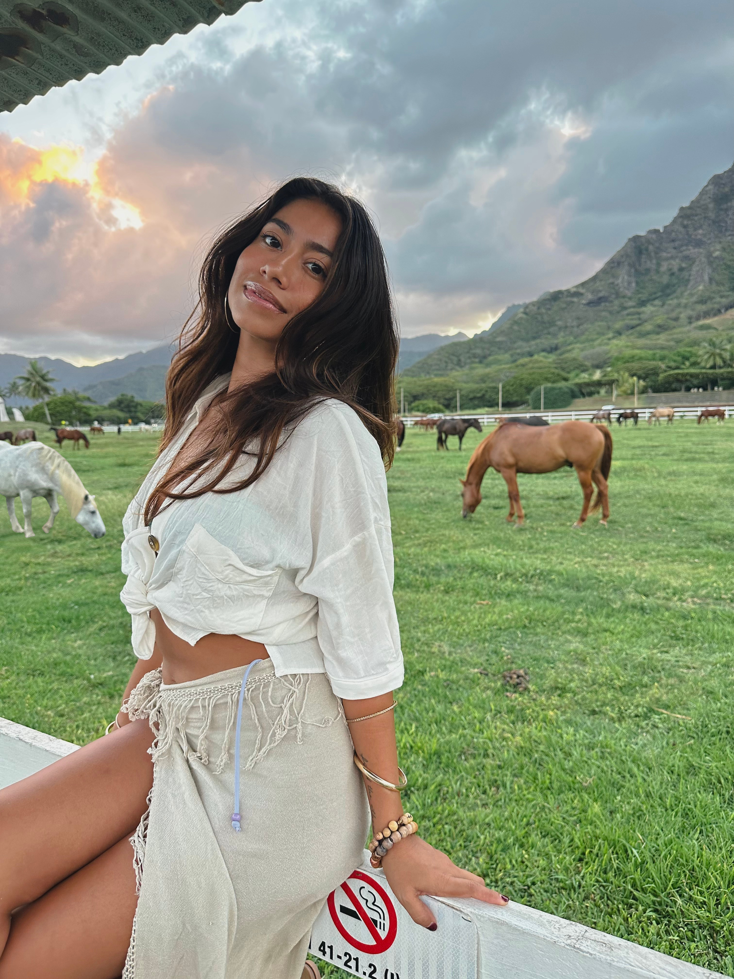 A woman with long dark hair, wearing a white cropped shirt and beige shorts, sitting outdoors at a horse farm with green fields and mountains in the background, during sunset with cloudy skies. Jenny Ochoa a singer on Oahu at Kualoa Ranch