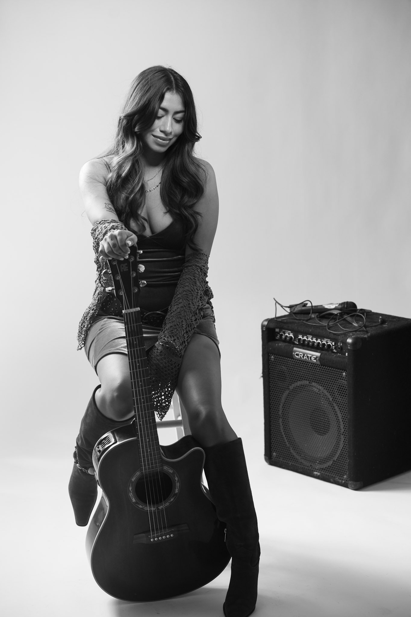 A woman with long wavy hair sitting on a stool, holding an acoustic guitar, with an amplifier and cables nearby, in a studio setting. Singers of Oahu. Live music on the island of Oahu