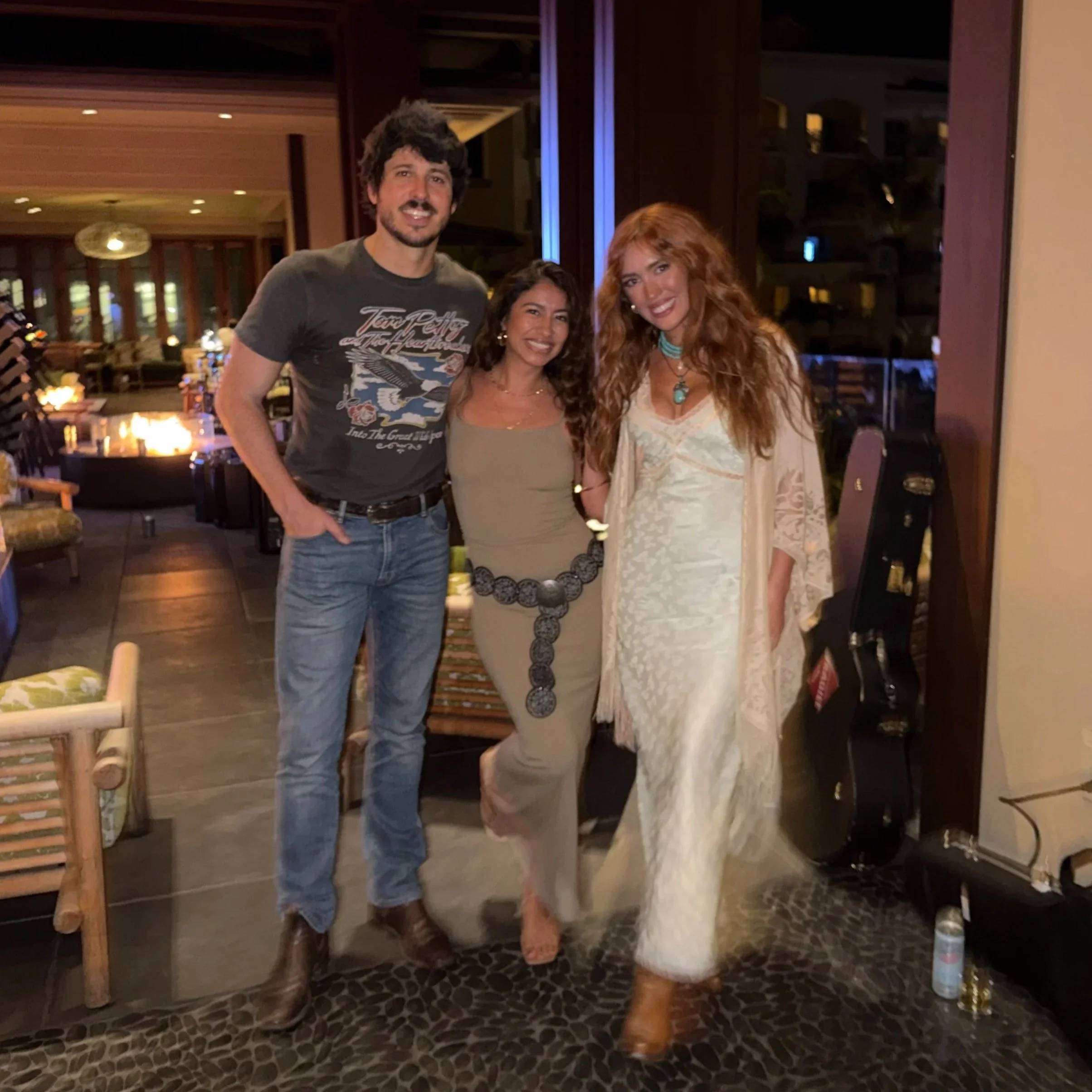 Three people standing together inside a warmly lit room, smiling at the camera, with a guitar in a case on the floor beside them. Laci Kaye Booth and Morgan Evans with Jenny Ochoa