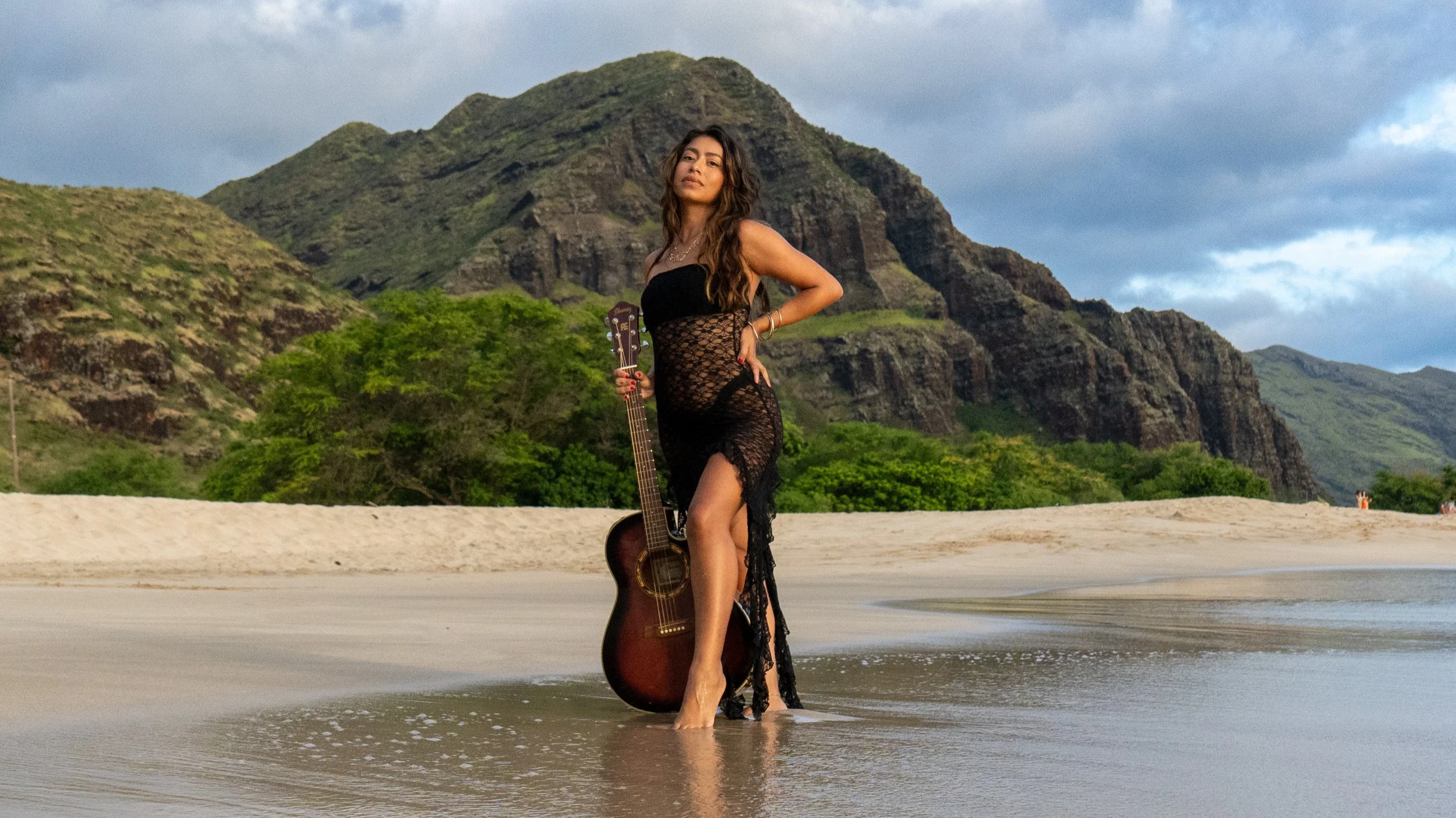 Woman in a black lace dress standing in shallow water on the beach holding a guitar with lush green mountains and cloudy sky in the background. Hawaii singer. Live music on Oahu.