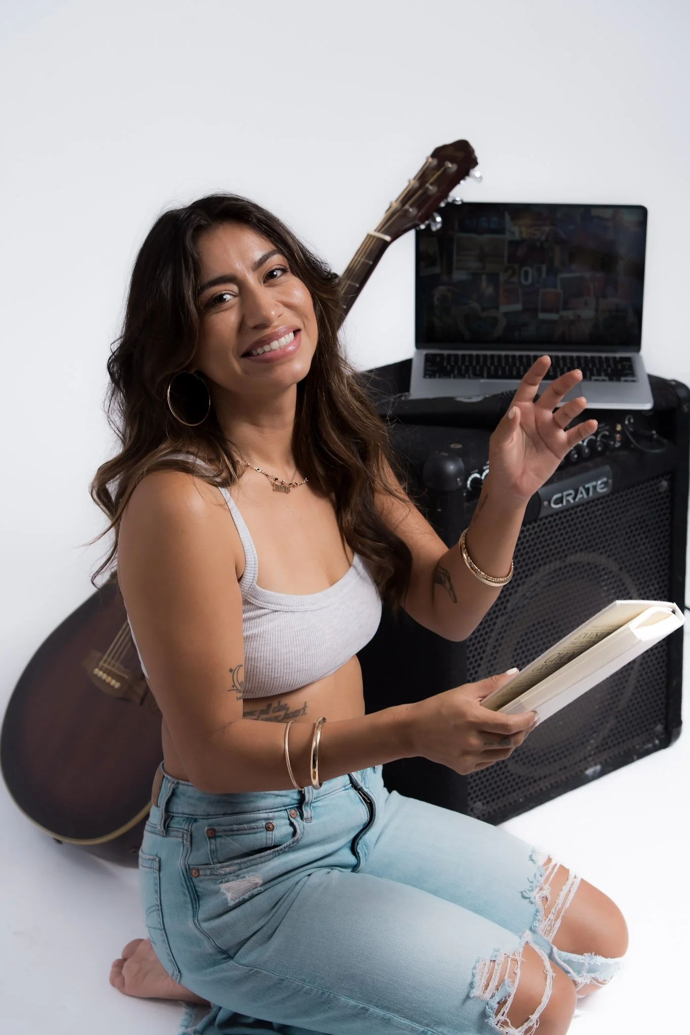 A woman with tattoos, wearing a gray tank top and ripped jeans, sitting on the floor with a guitar on her back, smiling, holding a notebook, with a laptop and amplifier behind her.