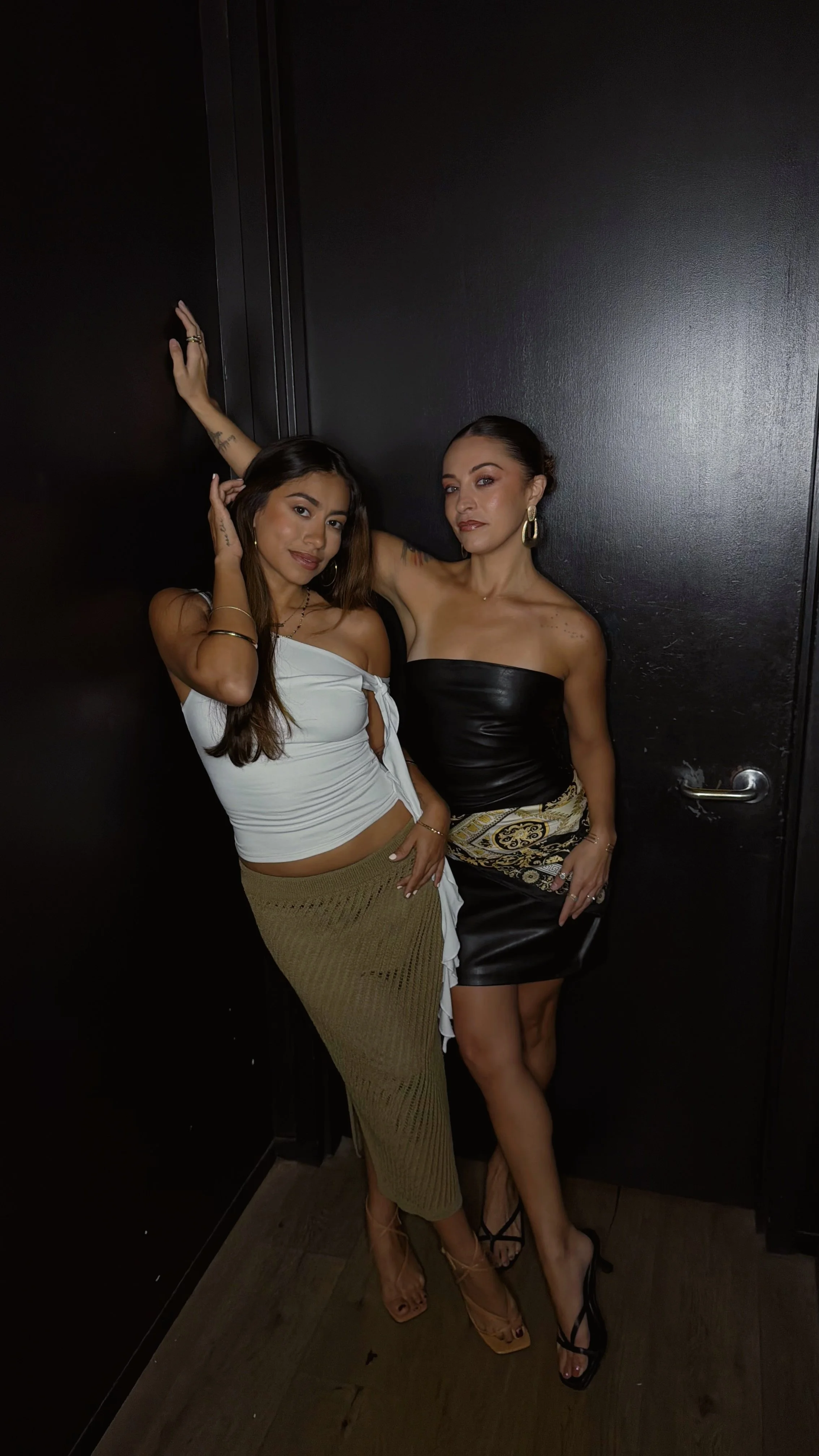 Two women posing together indoors, one wearing a white top and brown skirt, and the other wearing a black dress, both with jewelry and high-heeled shoes. Singer and make up artist on the island of Oahu