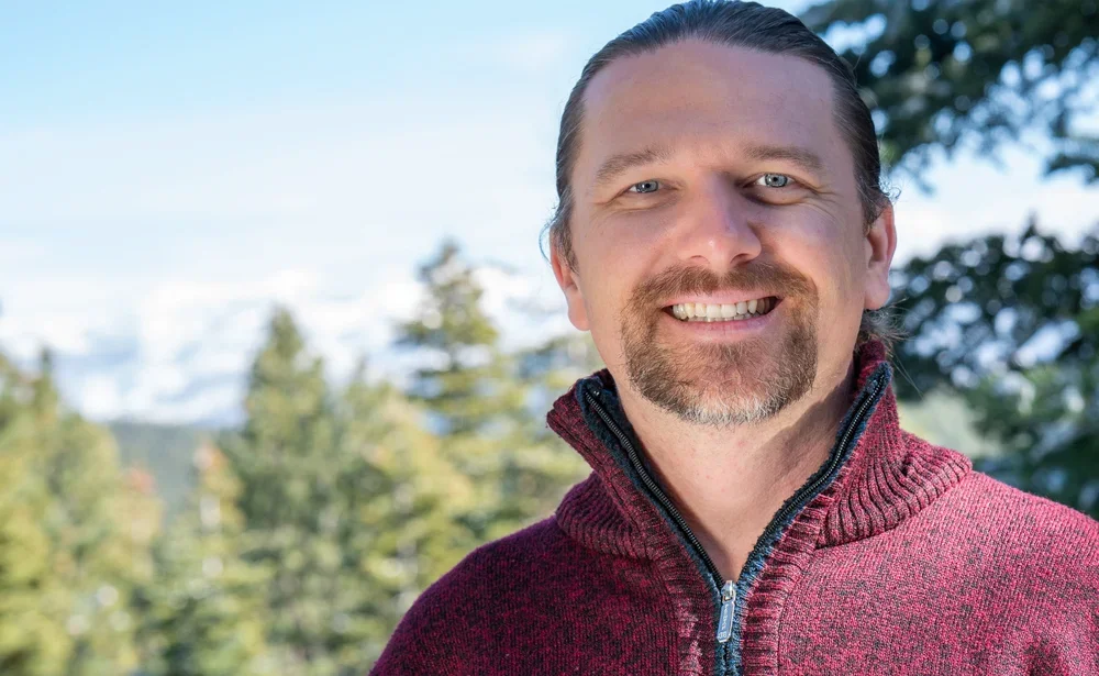 A man with a beard and mustache smiling outdoors near trees and a blue sky.