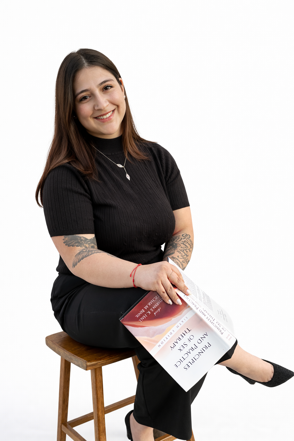 A woman with long brown hair, tattoos, and a nose piercing, sitting on a wooden stool and holding a magazine titled 'PANCHURCH PAKISTAN AND SEX THEORY' against a white background.