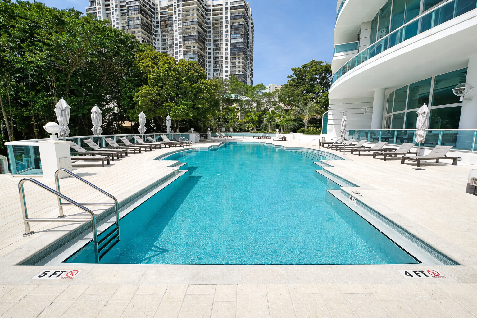 Empty outdoor hotel swimming pool with lounge chairs and umbrellas on a sunny day, surrounded by modern high-rise buildings and lush green trees.