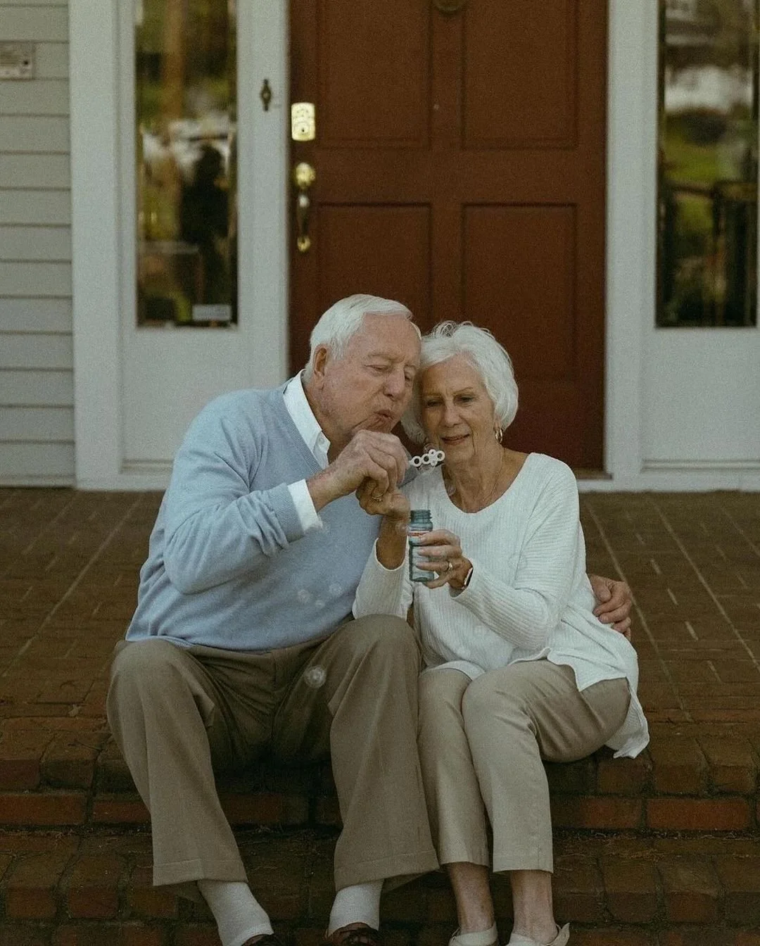 An elderly man and woman sitting on the front steps of a house, blowing bubbles together.