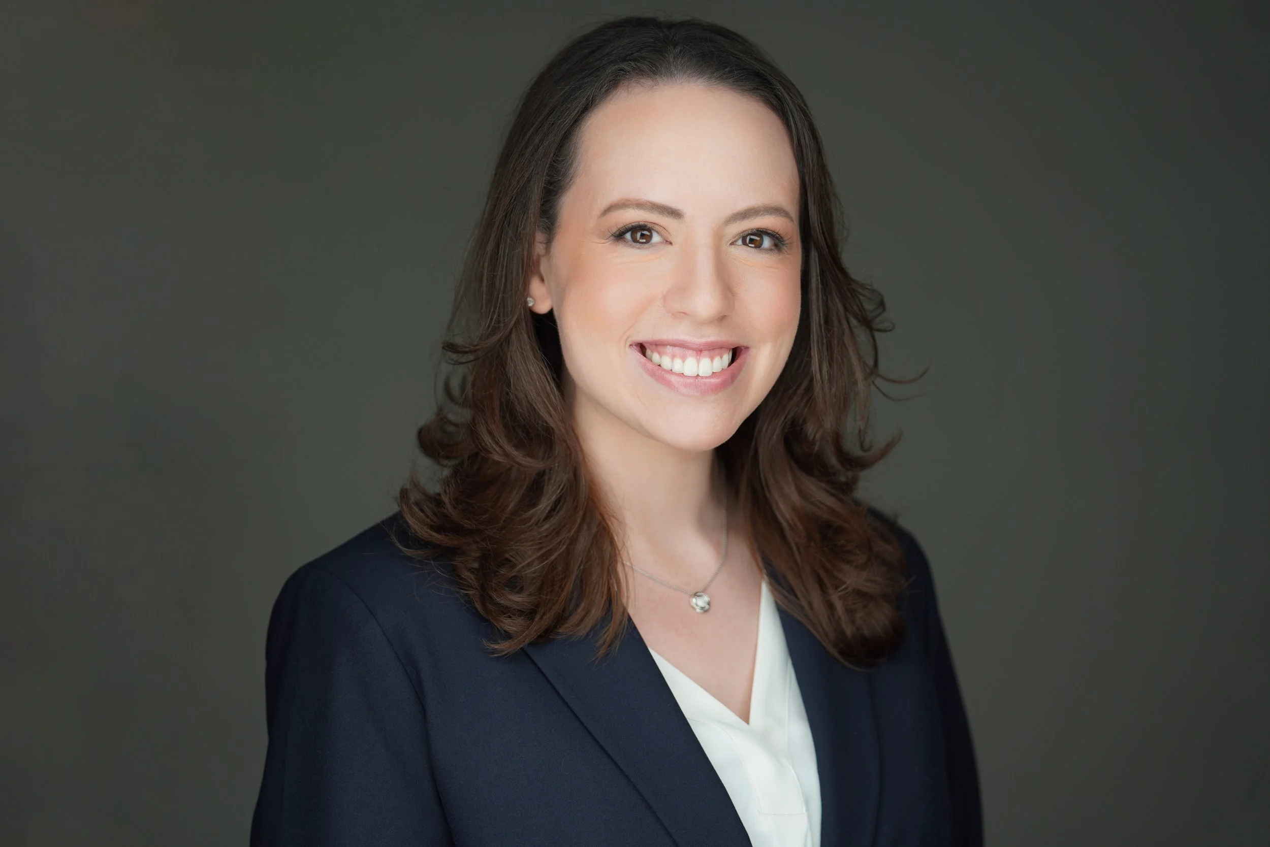 A woman with dark brown, wavy hair and a bright smile, wearing a navy blazer and a white top, against a gray background.