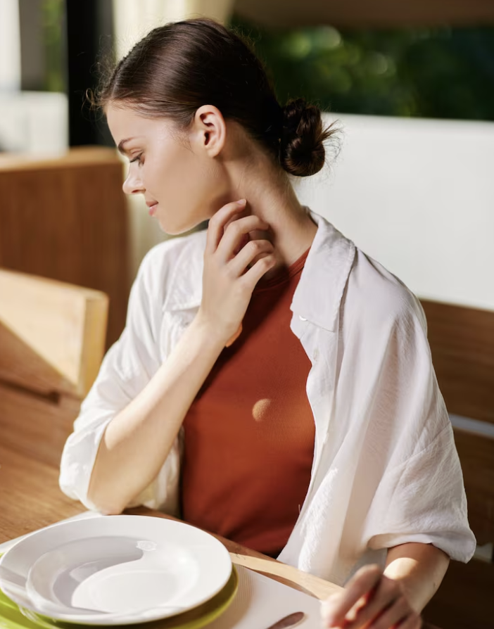A woman sitting at a table with a white plate, touching her neck and looking down thoughtfully, in a setting with natural light.