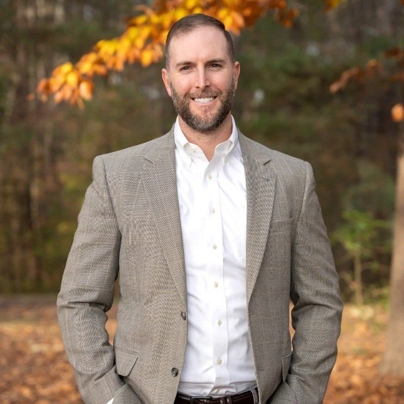 A man with a beard and short hair, wearing a light-colored blazer and white shirt, standing outdoors in front of trees with autumn leaves.