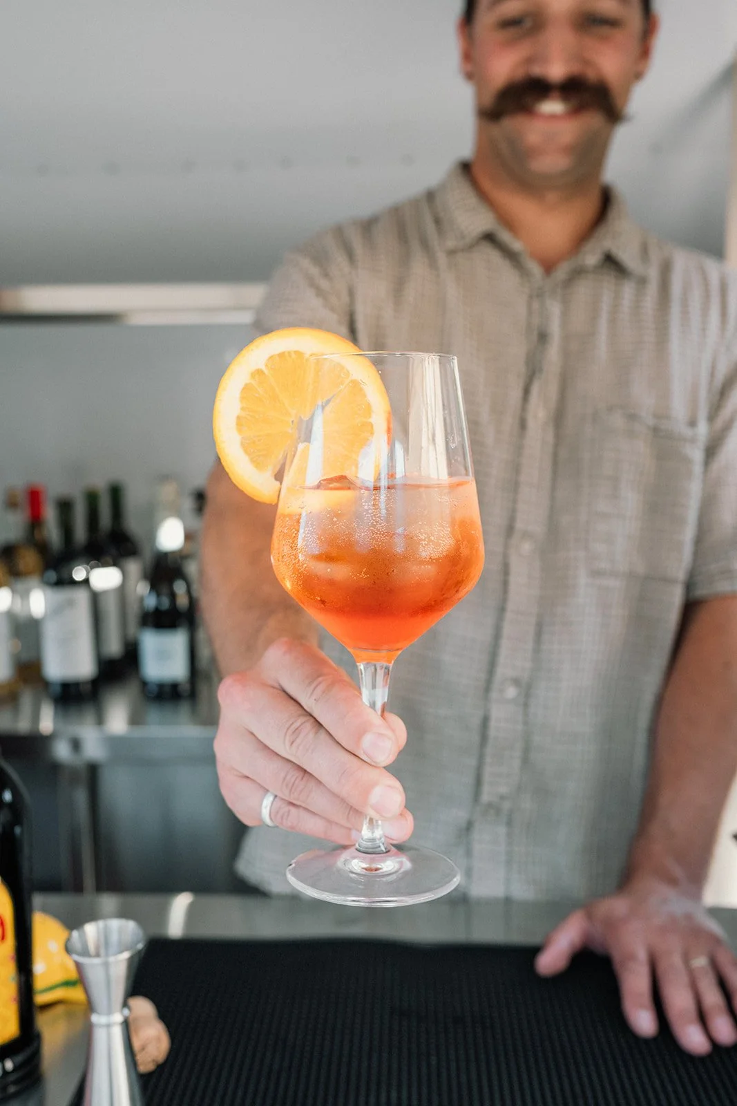 A man with a mustache and checked shirt holding a glass of orange-colored cocktail garnished with a lemon slice, with a bar background.