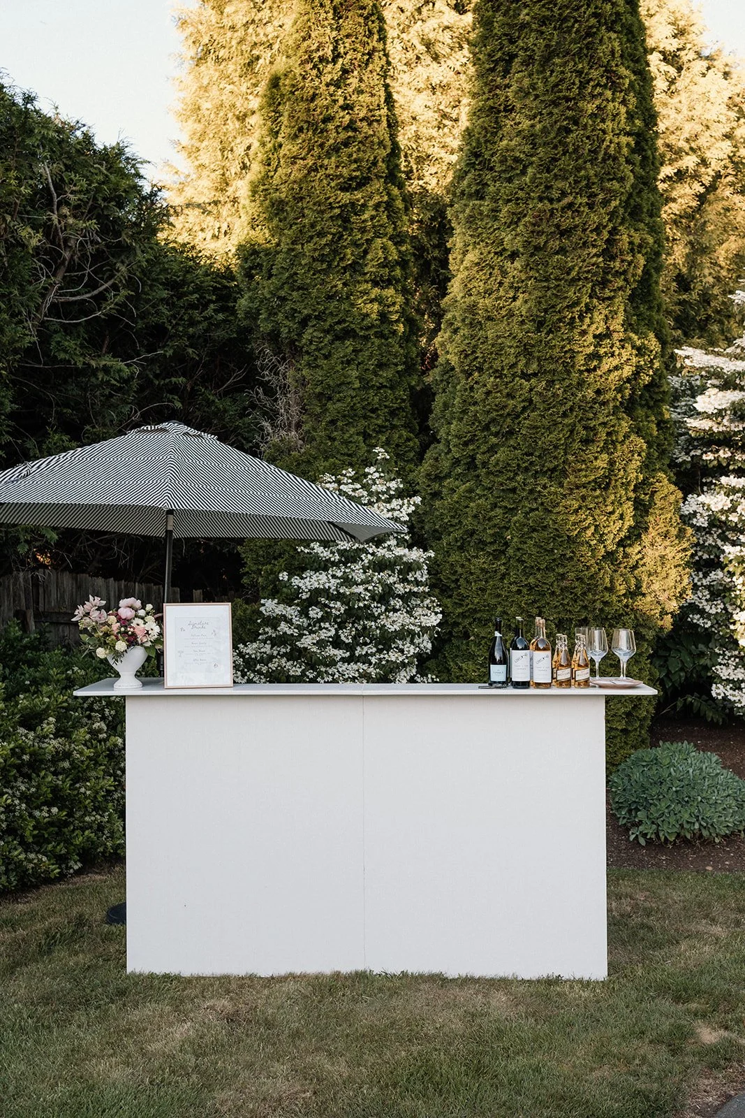 A white bar table set up outdoors with a striped patio umbrella, bottles of wine, wine glasses, and a flower arrangement in a vase, surrounded by lush green trees and shrubs.
