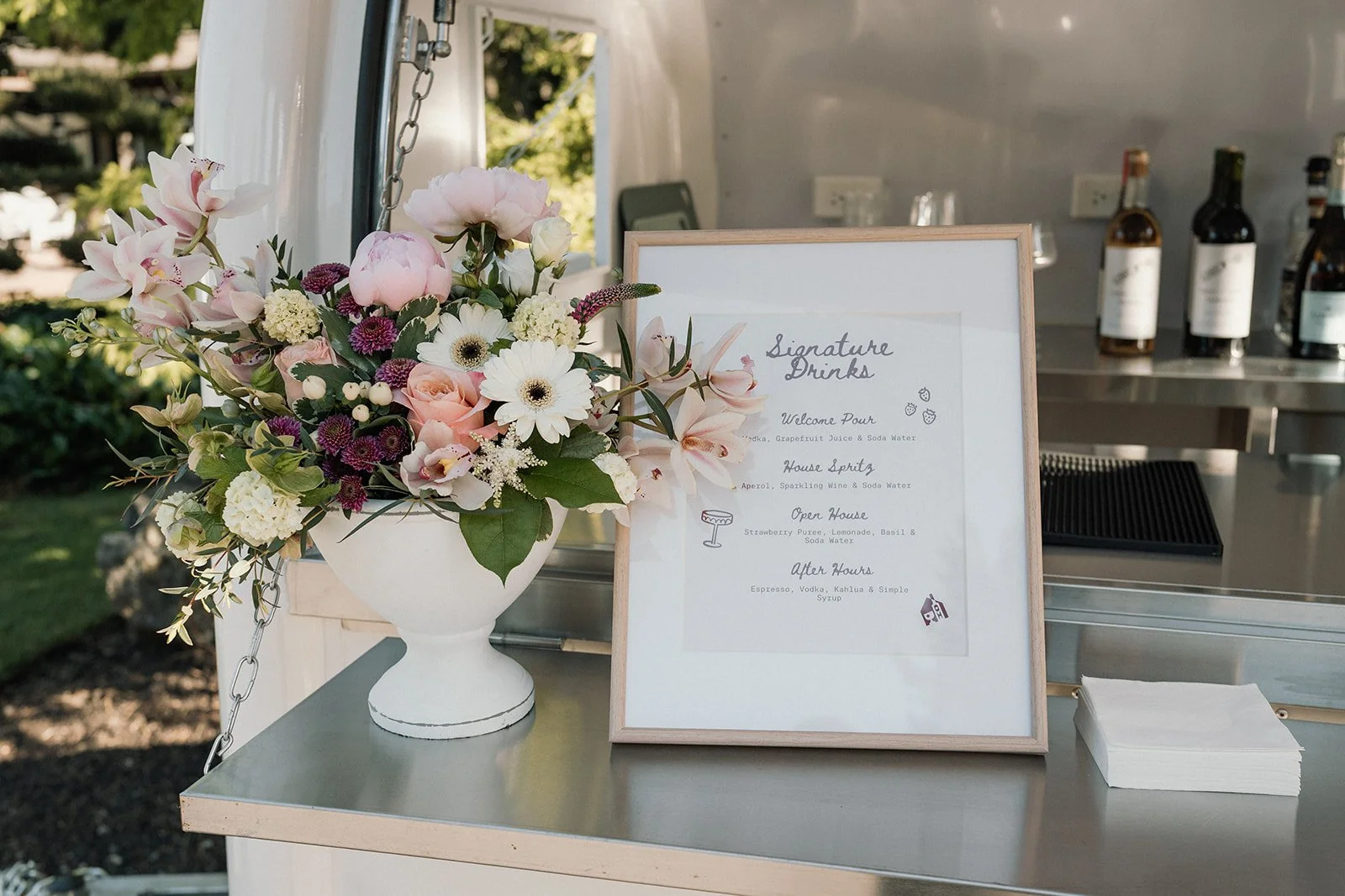 A floral arrangement in a white vase next to a framed sign listing drink options, situated on a stainless steel surface with bottles in the background.