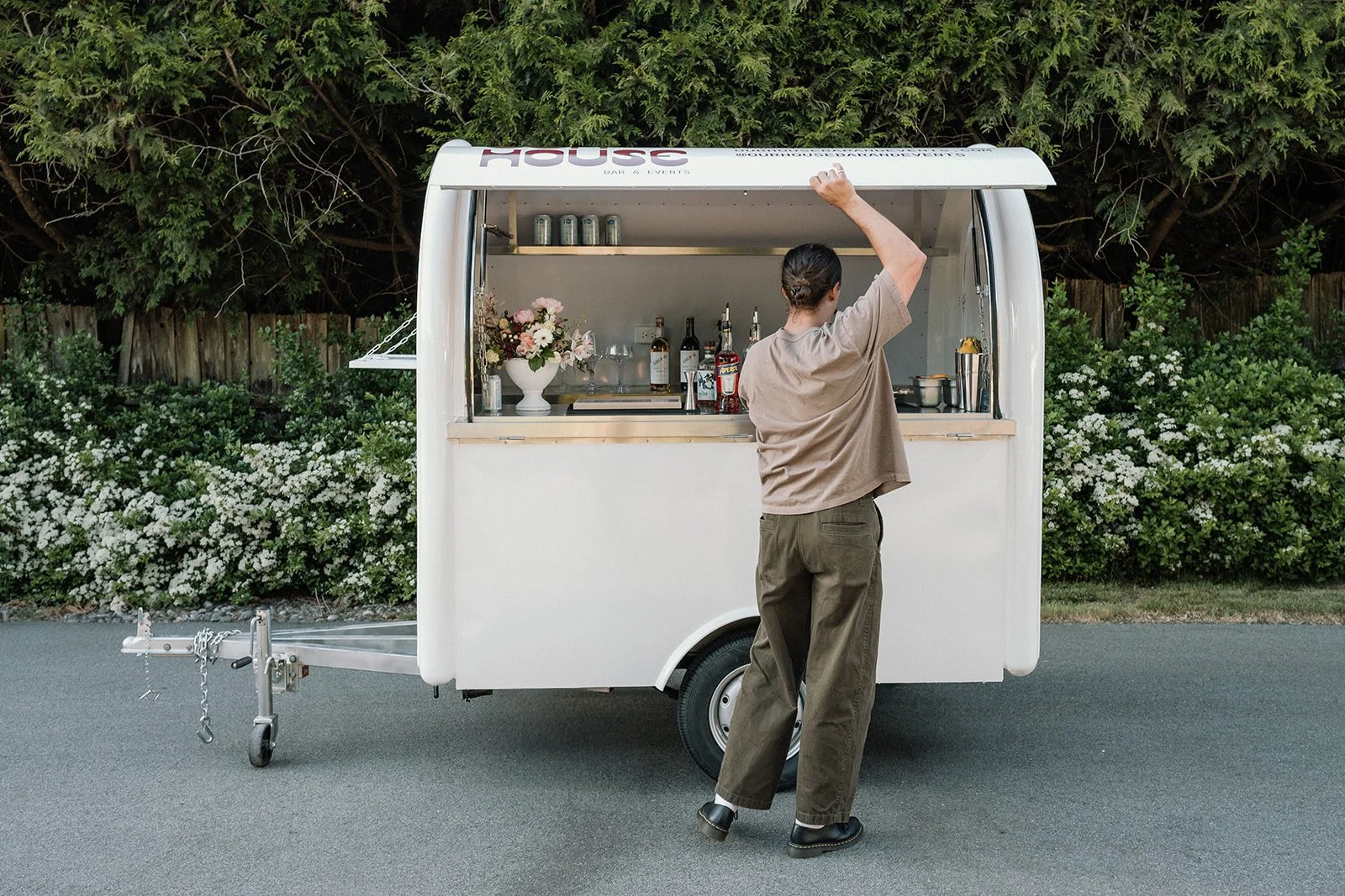 A person with brown hair tied back, wearing a beige shirt and loose brown pants, is standing in front of a small white mobile bar or food truck, reaching up to adjust the sign on the top. The truck is parked on a paved surface with green bushes and trees in the background.