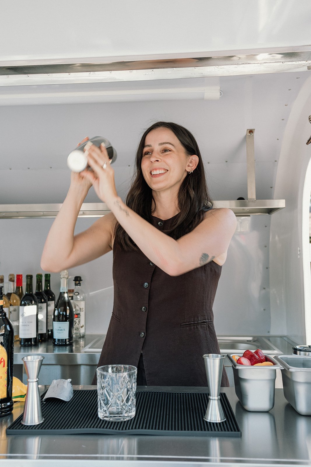 A woman smiling and shaking a cocktail shaker behind a bar counter with various bottles, strawberries, and bartending tools.