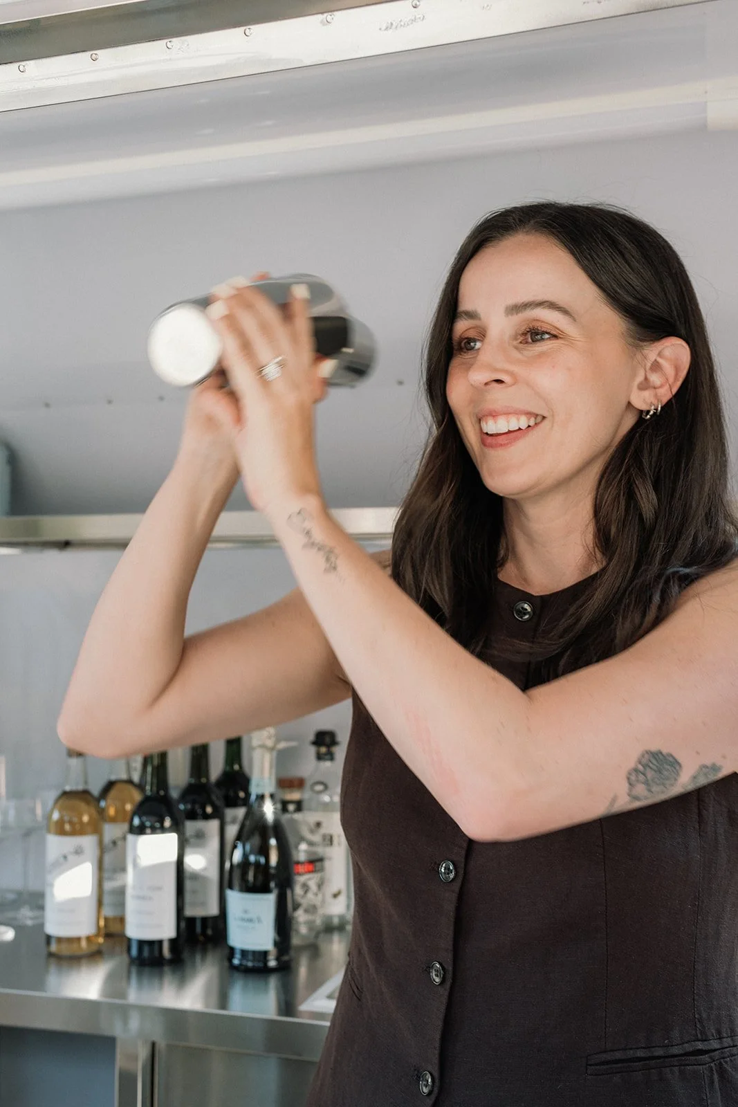 Woman with long dark hair, smiling, wearing a sleeveless black dress, holding a silver cocktail shaker, with a tattoo on her left forearm, in front of a bar with bottles of wine and spirits.