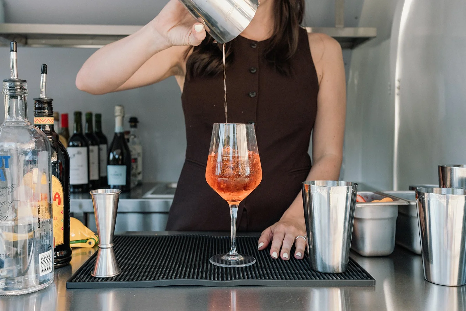 A woman wearing a sleeveless brown dress pouring a pink cocktail into a glass at a bar, surrounded by liquor bottles, cocktail utensils, and bar tools.