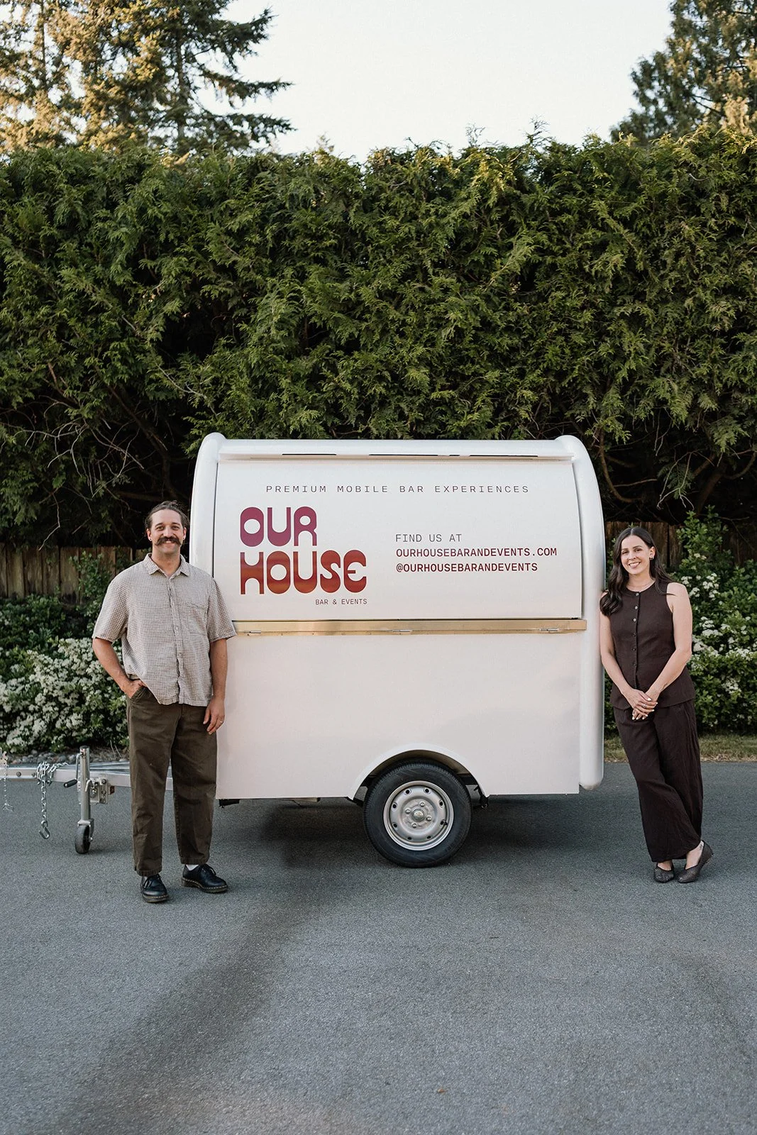 Man with mustache and woman standing beside a mobile bar trailer outdoors. The trailer has signs promoting Our House Bar & Events and a website. The background features trees and bushes.